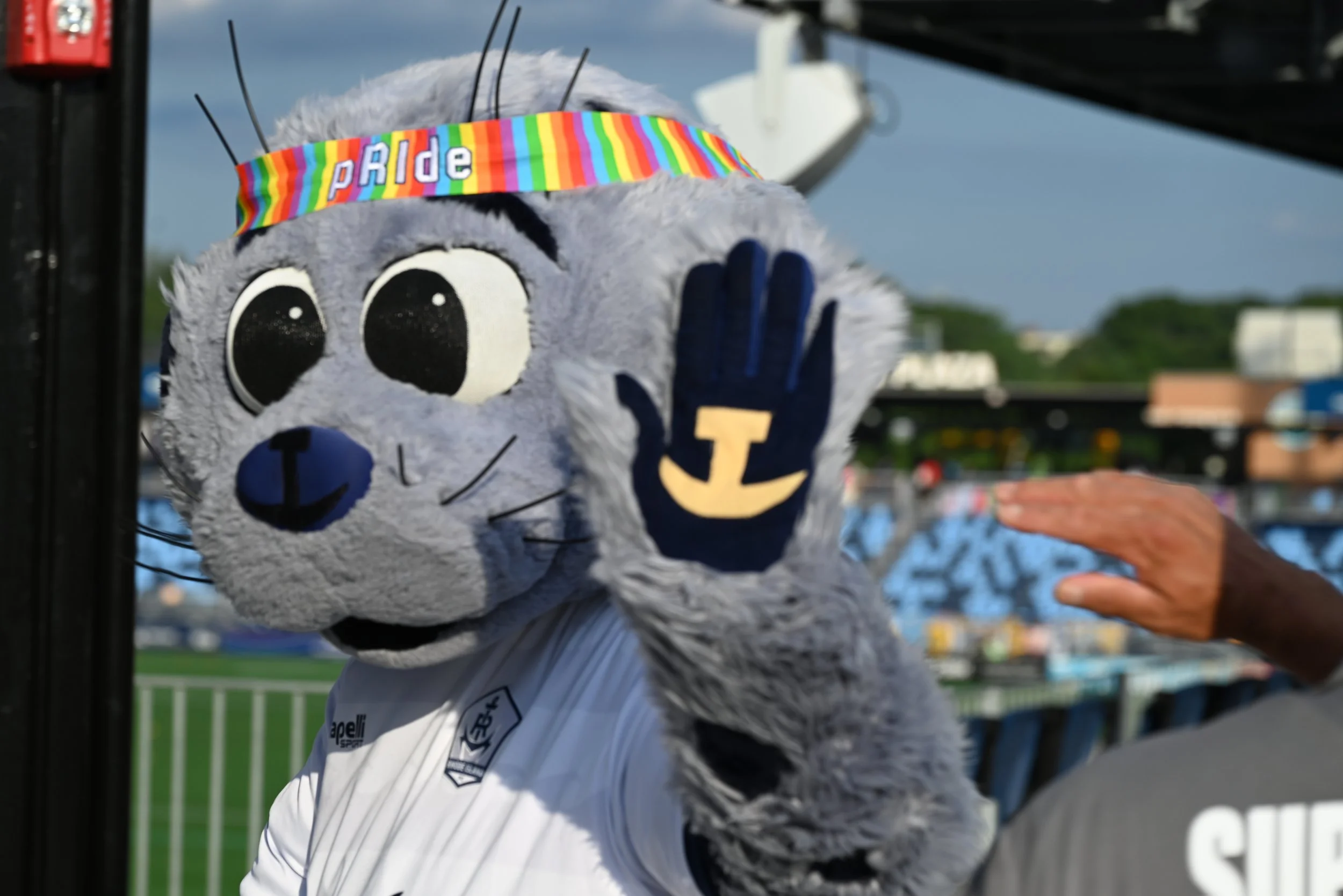 A person dressed in a mascot costume resembling a gray cat with large eyes, wearing a rainbow-colored headband with the word 'PRIDE' on it, waving at the camera during a parade or outdoor event.