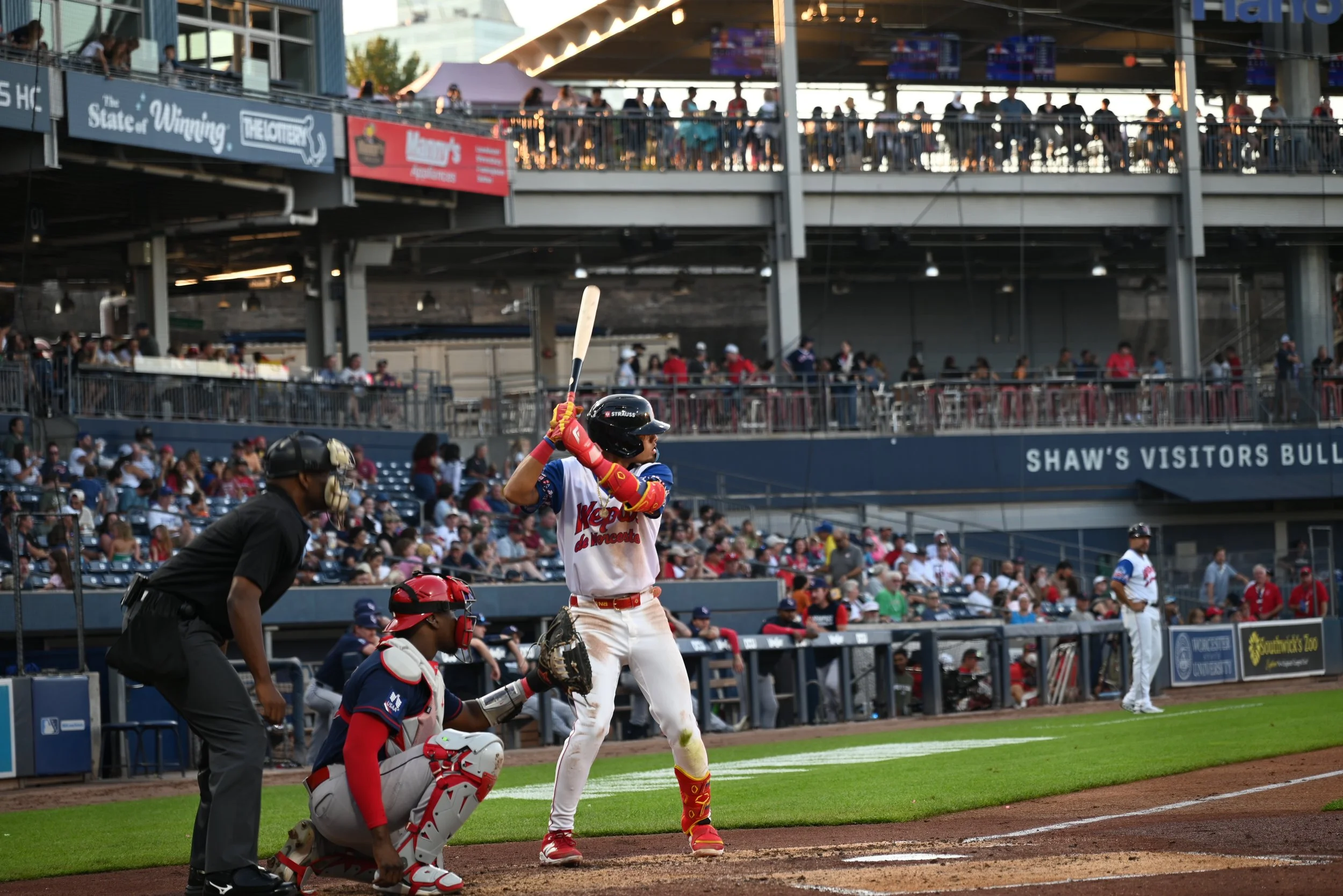 A baseball player in a white uniform with red and yellow accents stands at home plate holding a bat, ready to swing. An umpire and catcher are positioned behind him, and there are spectators in the background in a stadium.