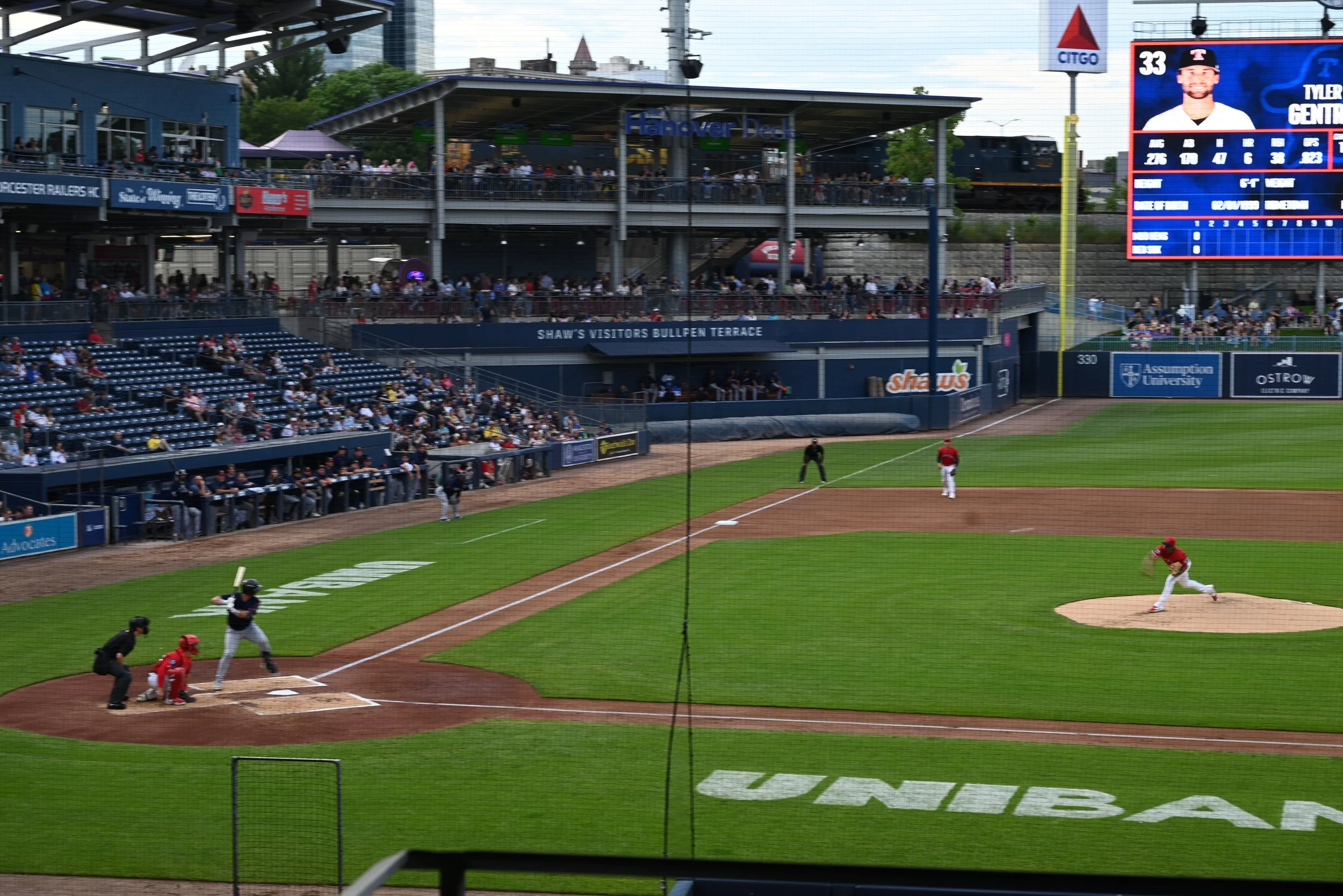 A baseball game in progress at a stadium with spectators in the stands and on the terraces. A batter is running towards first base, while a player in a red uniform fields the ball. The scoreboard shows player information and team scores.