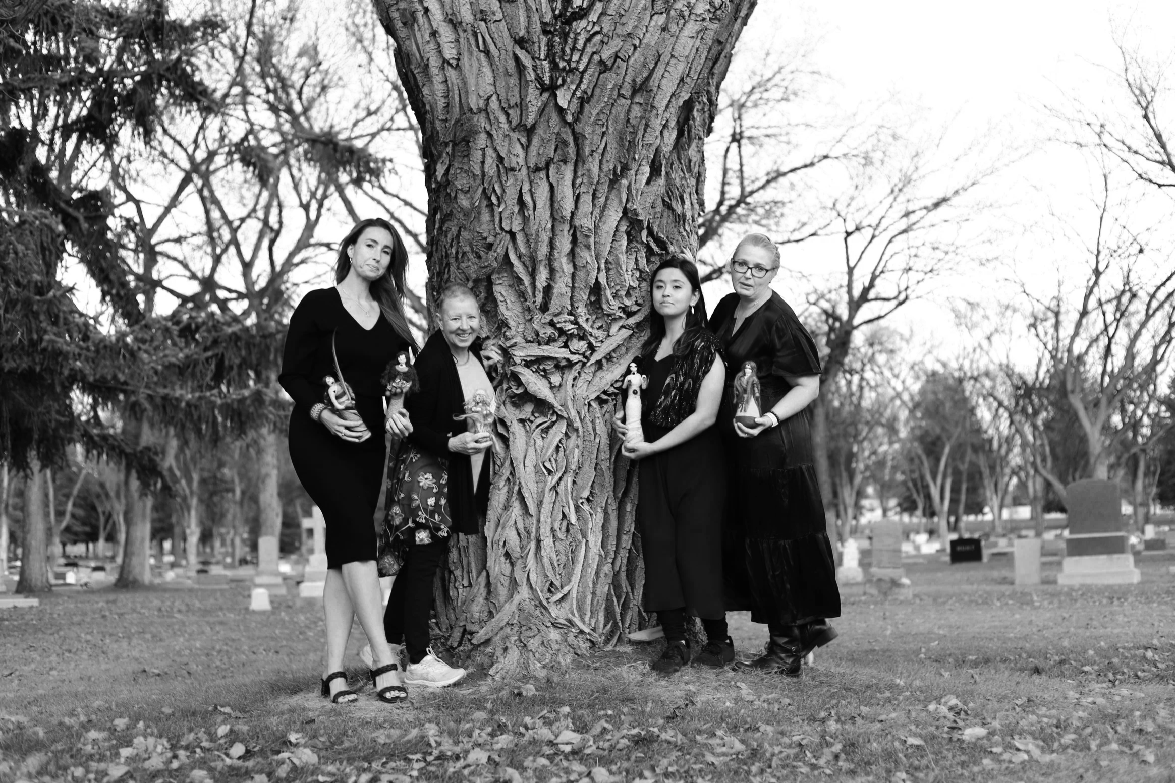 A black-and-white photo of four women standing outdoors around a large tree trunk in a park, holding various objects, with leafless trees and gravestones visible in the background.