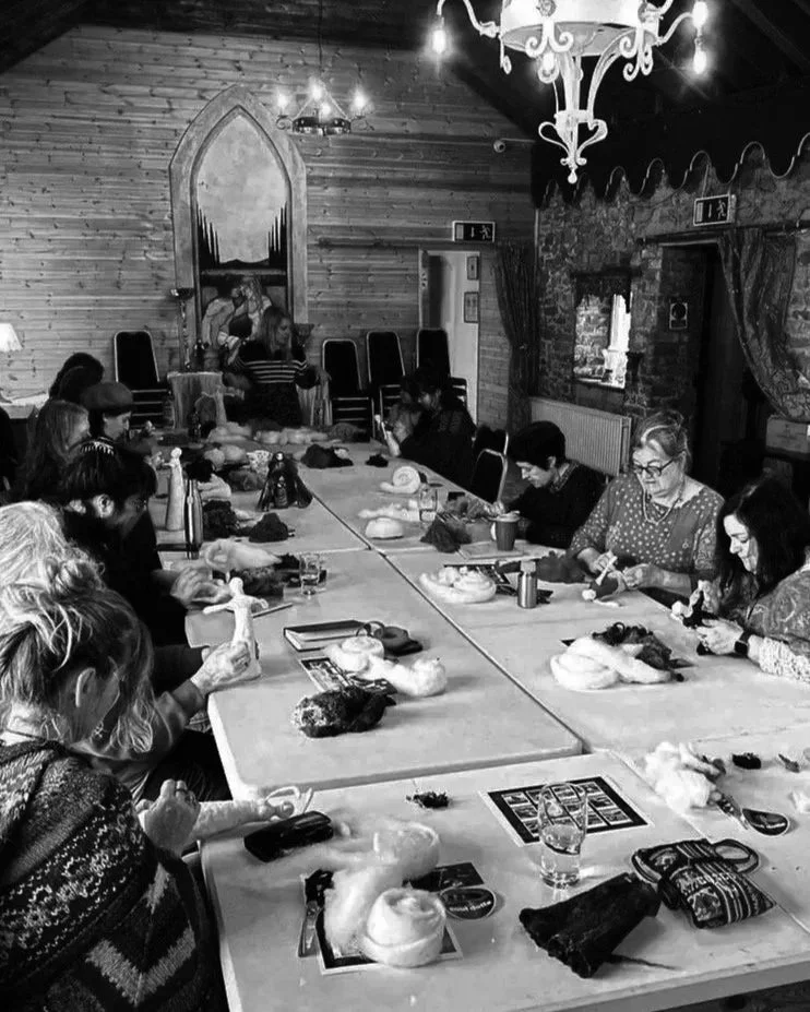 People sitting around a long table in a rustic room, knitting or crafting, with yarn and craft supplies on the table.