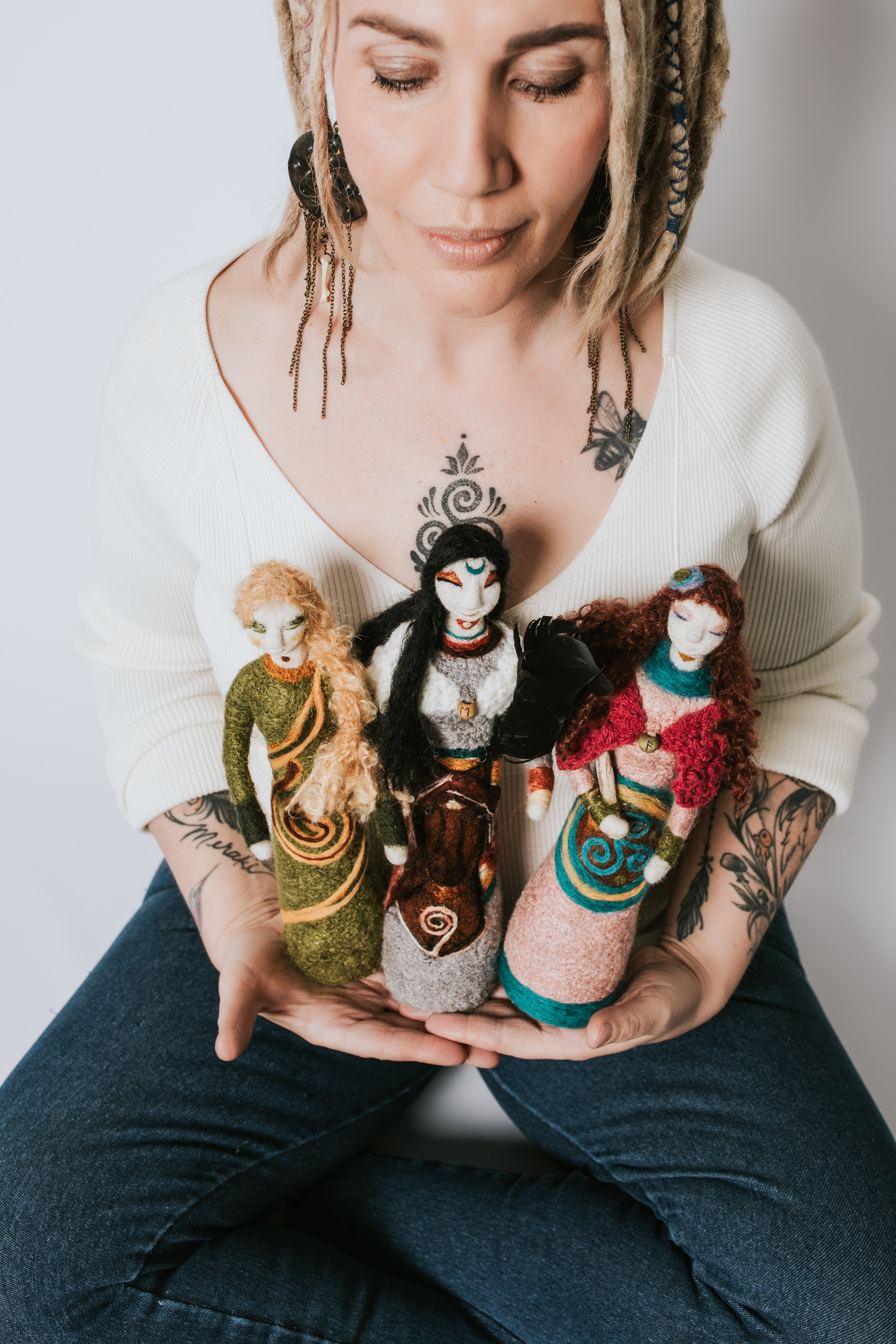 A woman with light skin, tattoos, and dreadlocks, wearing a white top, holding a collection of colorful felt dolls.