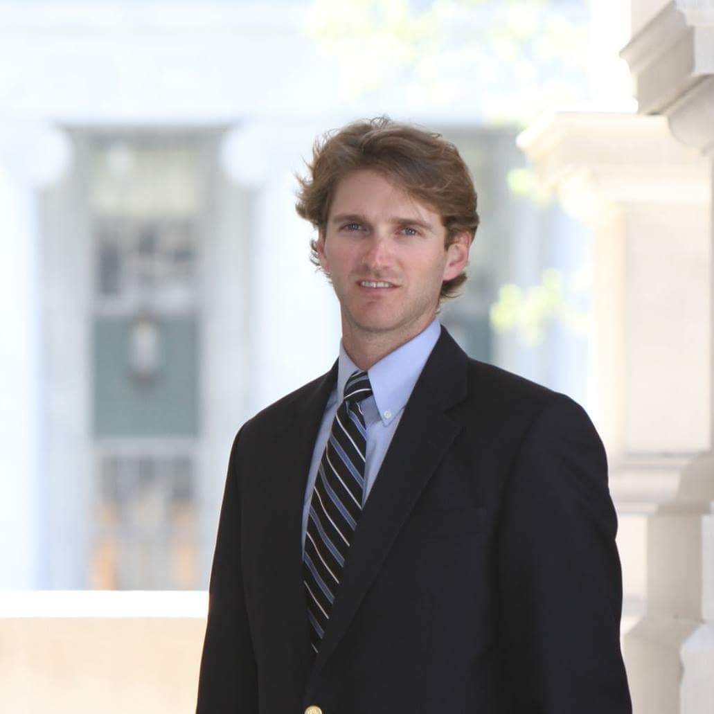 A young man in a black suit with a blue shirt and striped tie standing outdoors in front of out-of-focus columns or pillars.