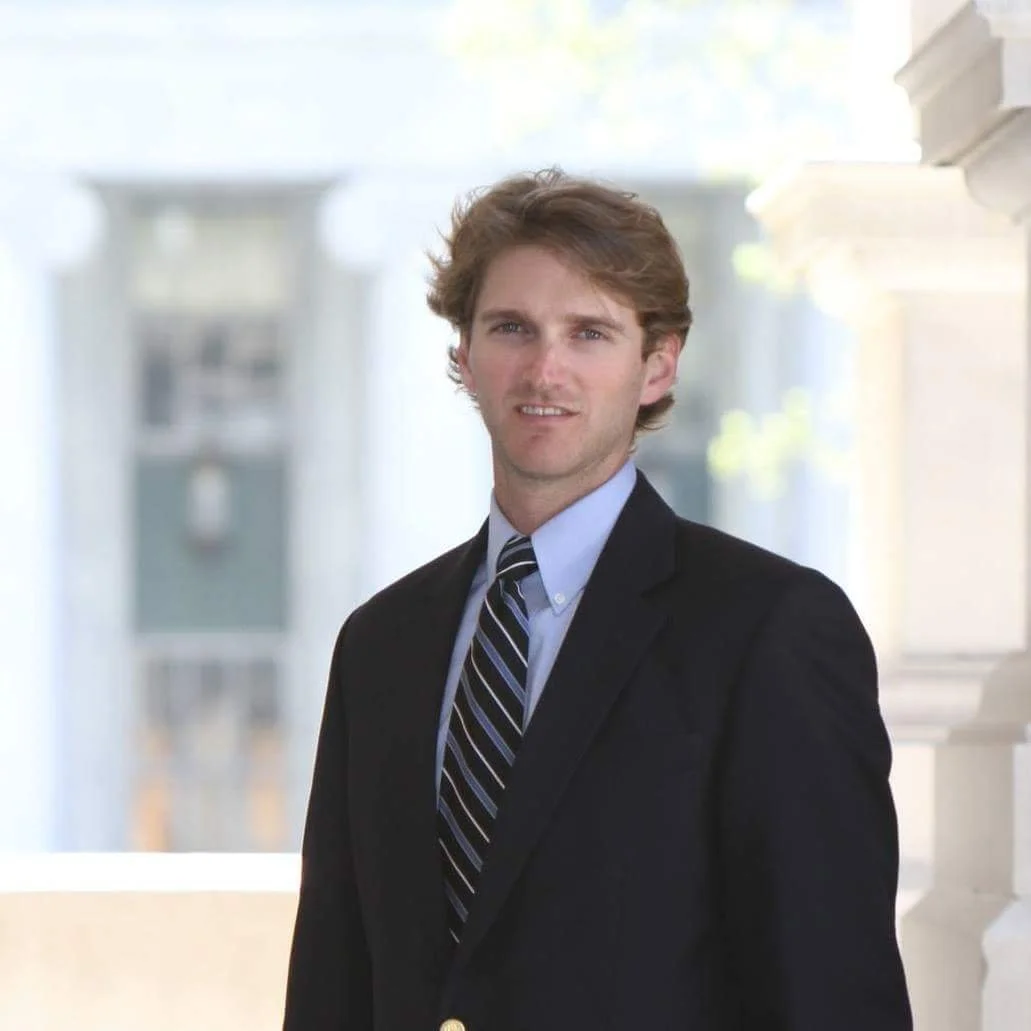 A young man with light brown hair wearing a dark suit, white shirt, and striped tie standing outdoors in front of a blurred background of a building with white columns.