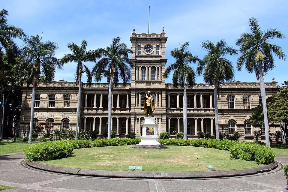 Historical government building with clock tower, surrounded by palm trees and a statue in front, in a tropical setting.
