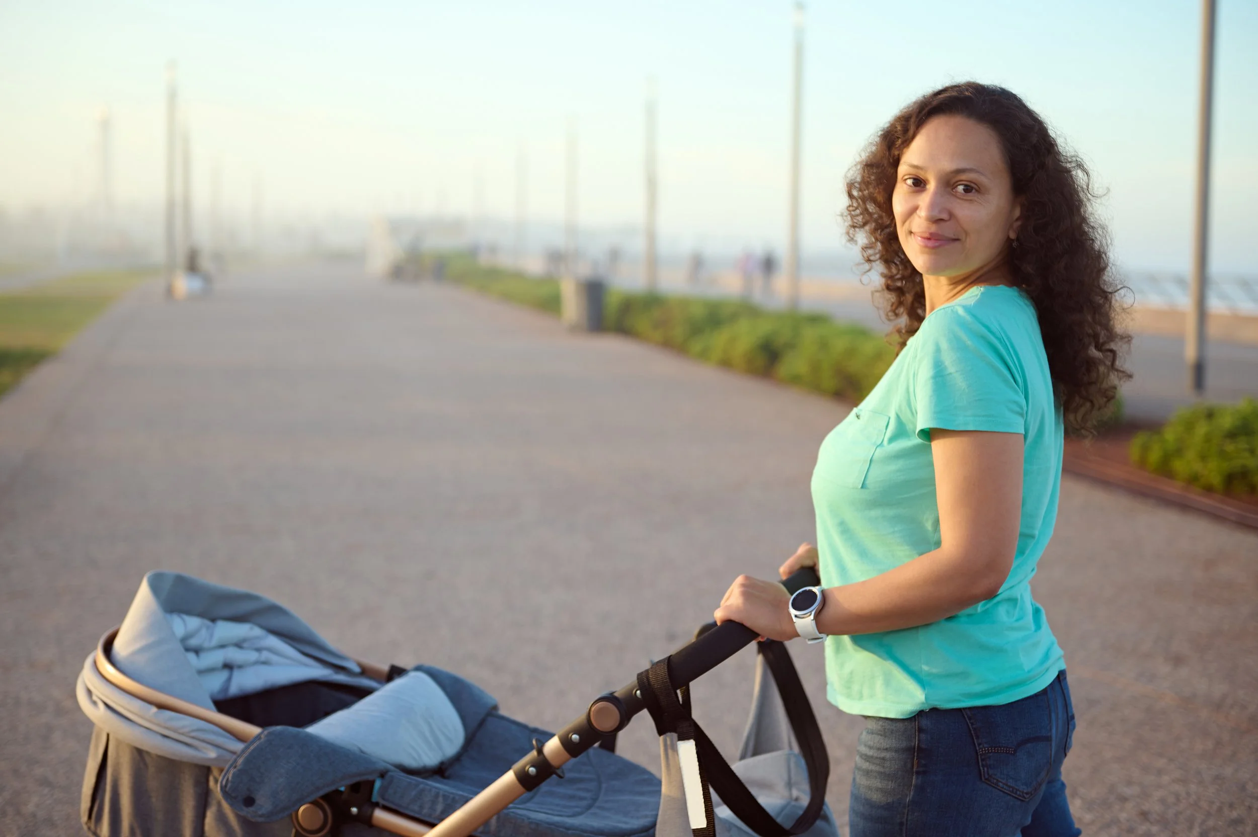 A woman or mom stands outdoors on a paved walkway with a baby stroller. The background includes a waterfront, green bushes, and a few distant people on a foggy or misty day. The woman looks happy in her role.