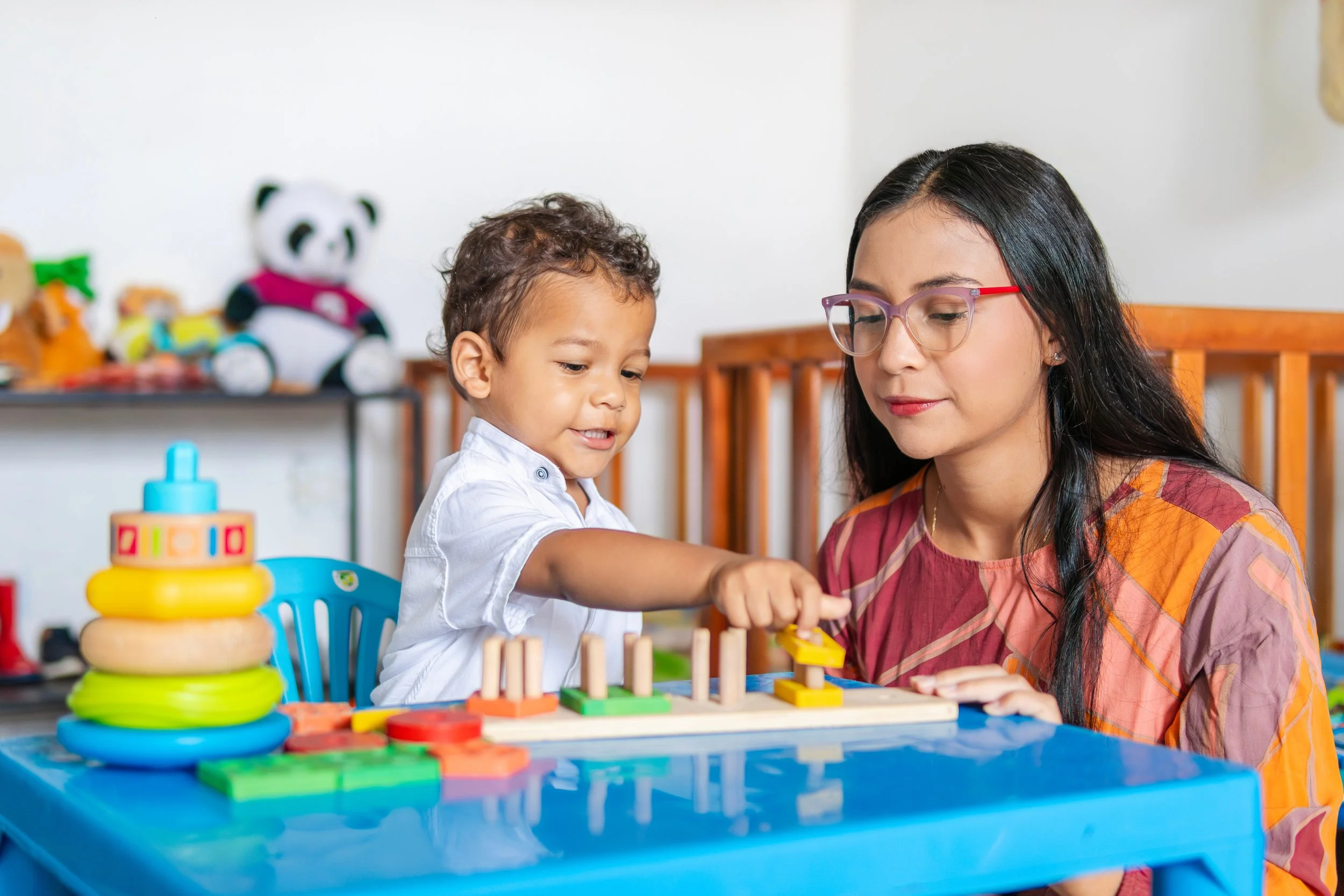 A young boy and a caregiver with long dark hair and glasses play a wooden pegboard game at a blue table.