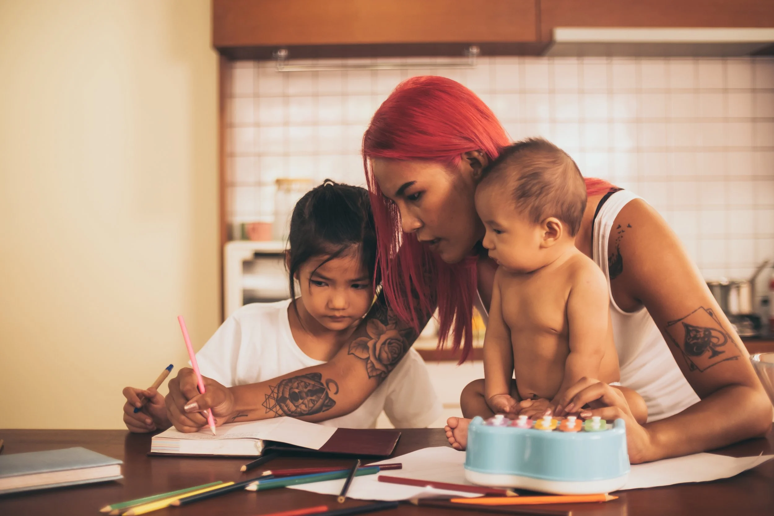 An Asian caregiver or mother with red hair and tattoos is helping two young children with drawing and coloring at a table in a kitchen.