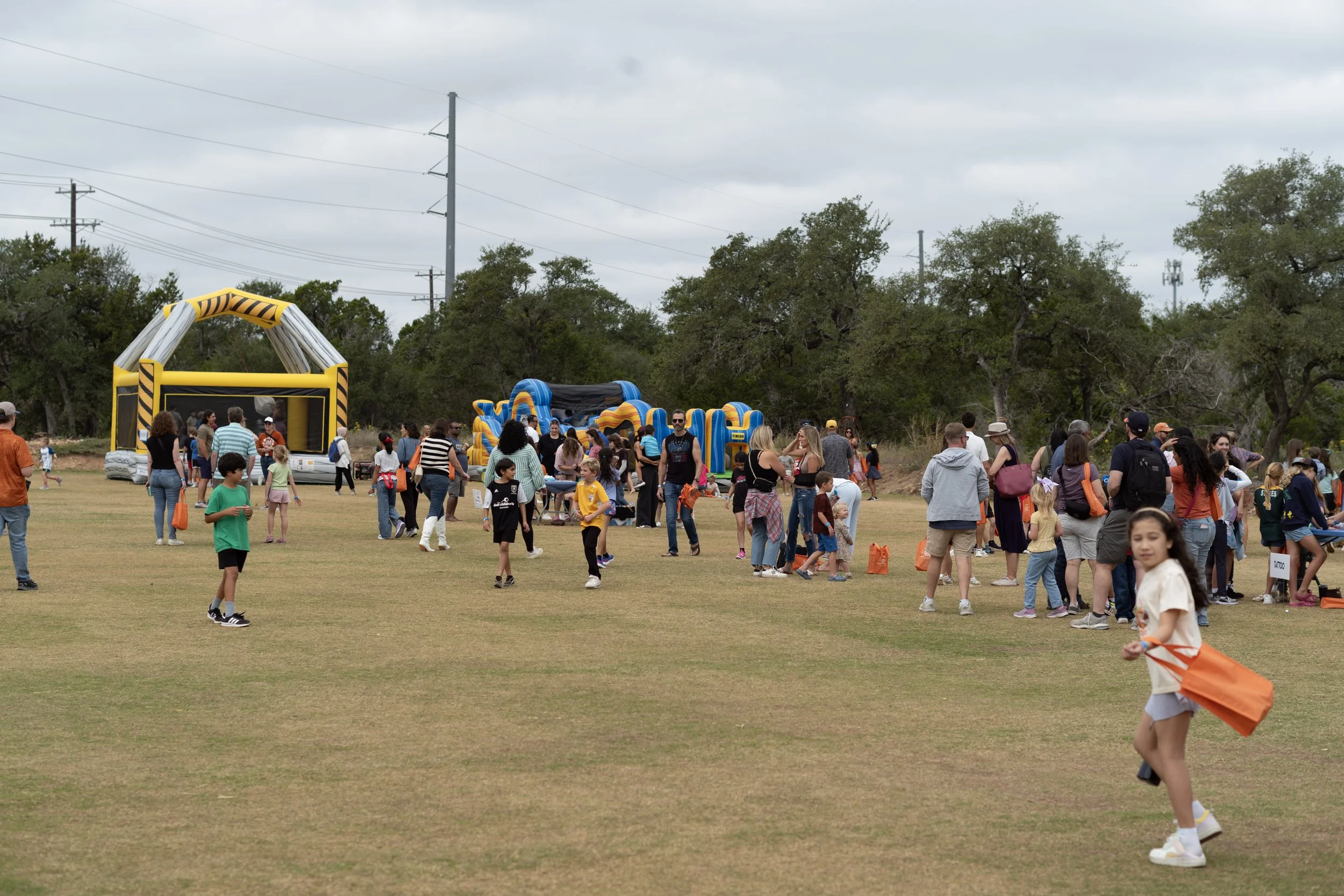A crowded outdoor event in a grassy field with inflatable bounce houses and many people, including children and adults, participating in activities on a cloudy day.