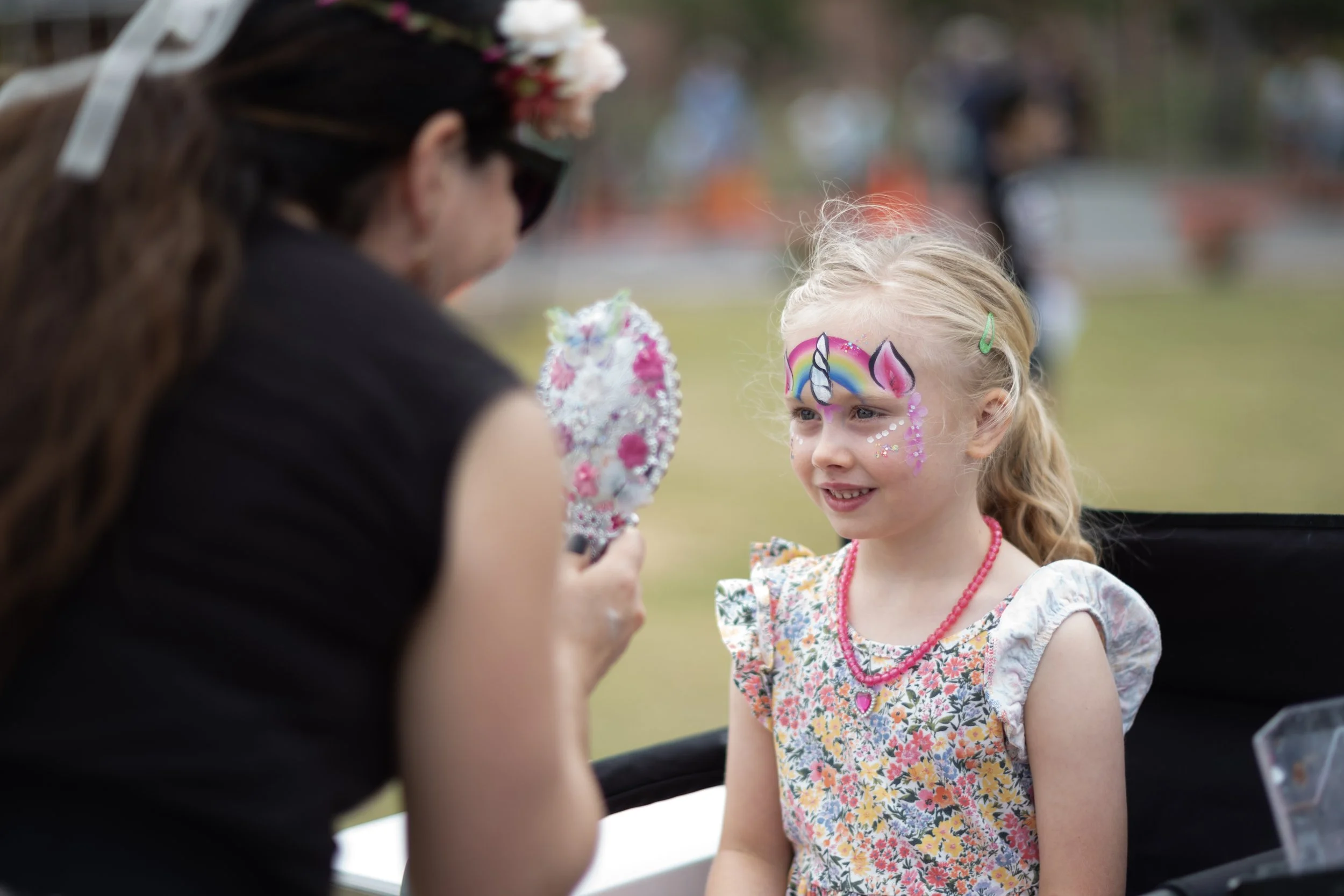 A young girl with face paint of a rainbow, unicorn, and pink accents is getting her face painted by an adult at an outdoor event. The girl is wearing a floral dress and pink necklace, and she has a green hair clip. The adult is holding a paintbrush a