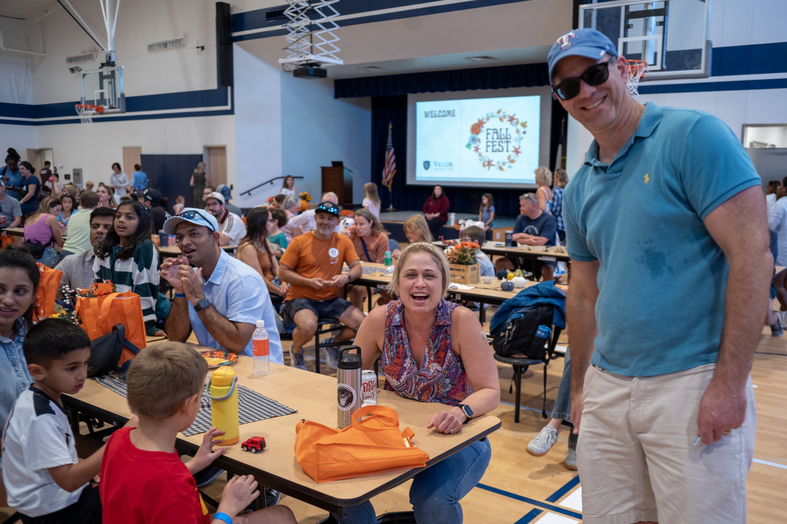 A group of people, including children and adults, gathered in a gymnasium for a fall festival event, with tables, decorations, and a stage displaying a 'Fall Fest' banner in the background.