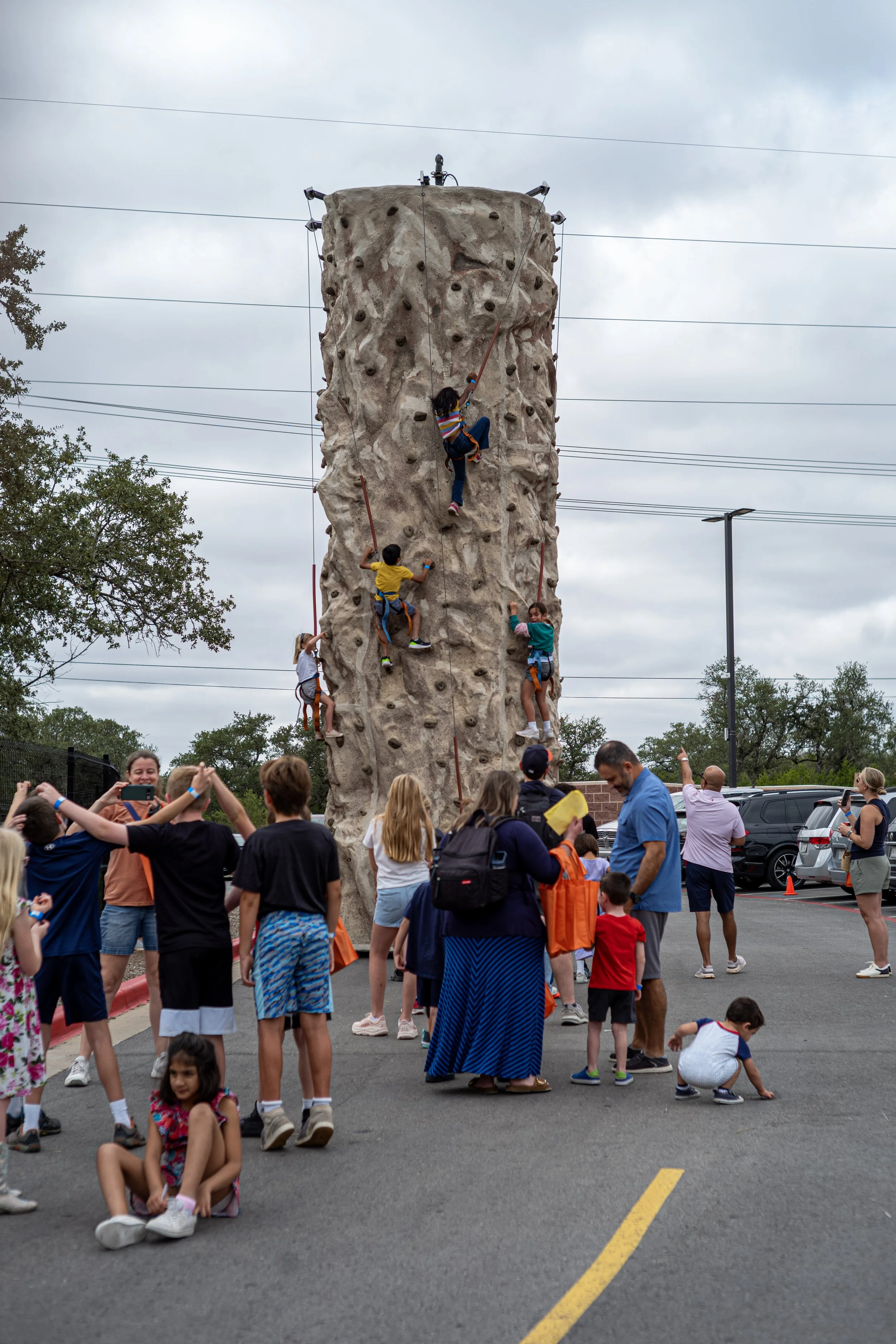 Children and adults gathered outside, watching or waiting near a large outdoor climbing wall with kids climbing it.