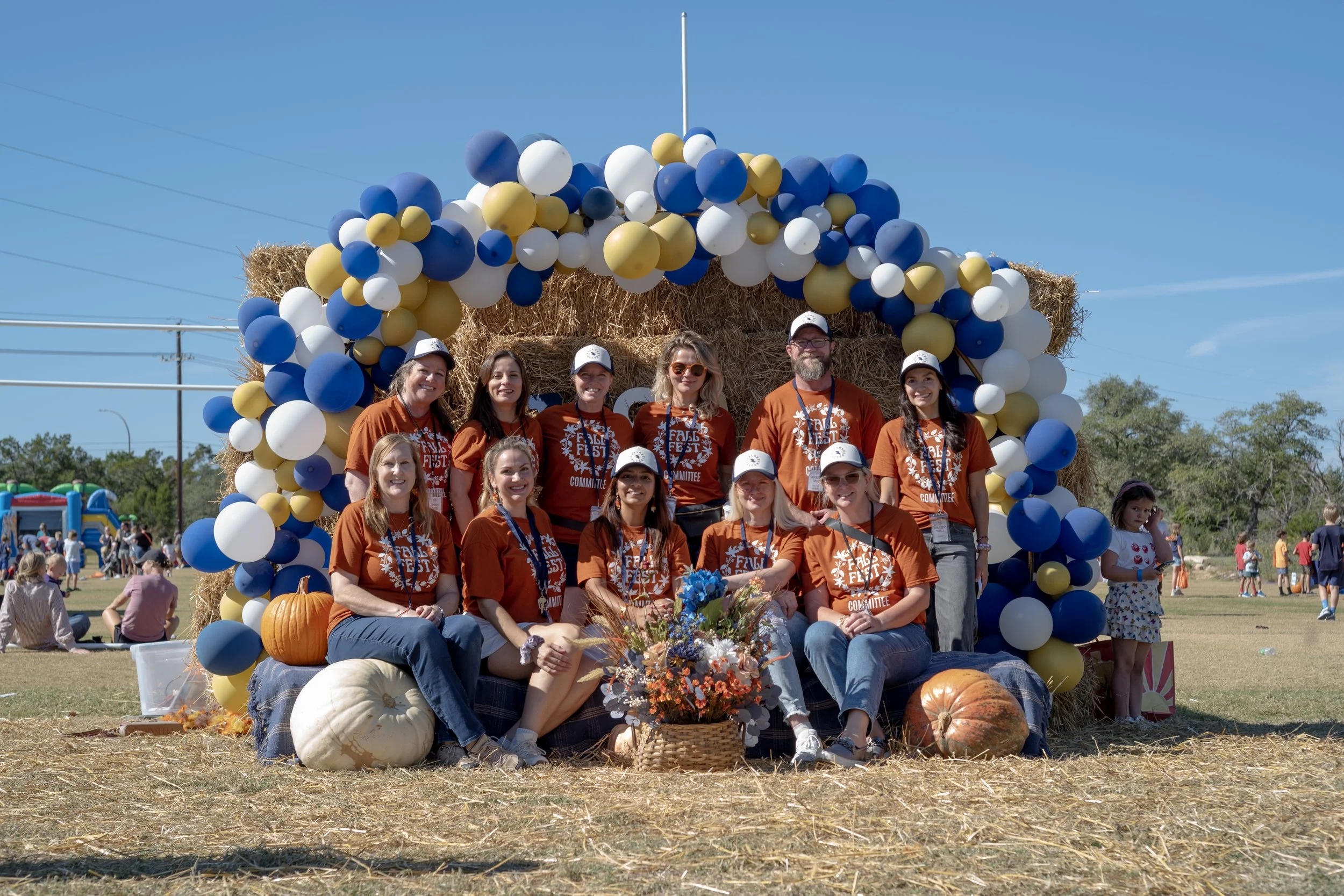 A group of people wearing matching orange shirts with white text, gathered in front of a straw bale arch decorated with blue, white, and yellow balloons at an outdoor fall festival. Fall pumpkins and flowers are at their feet, and other festival attractions and children are visible in the background under a clear blue sky.