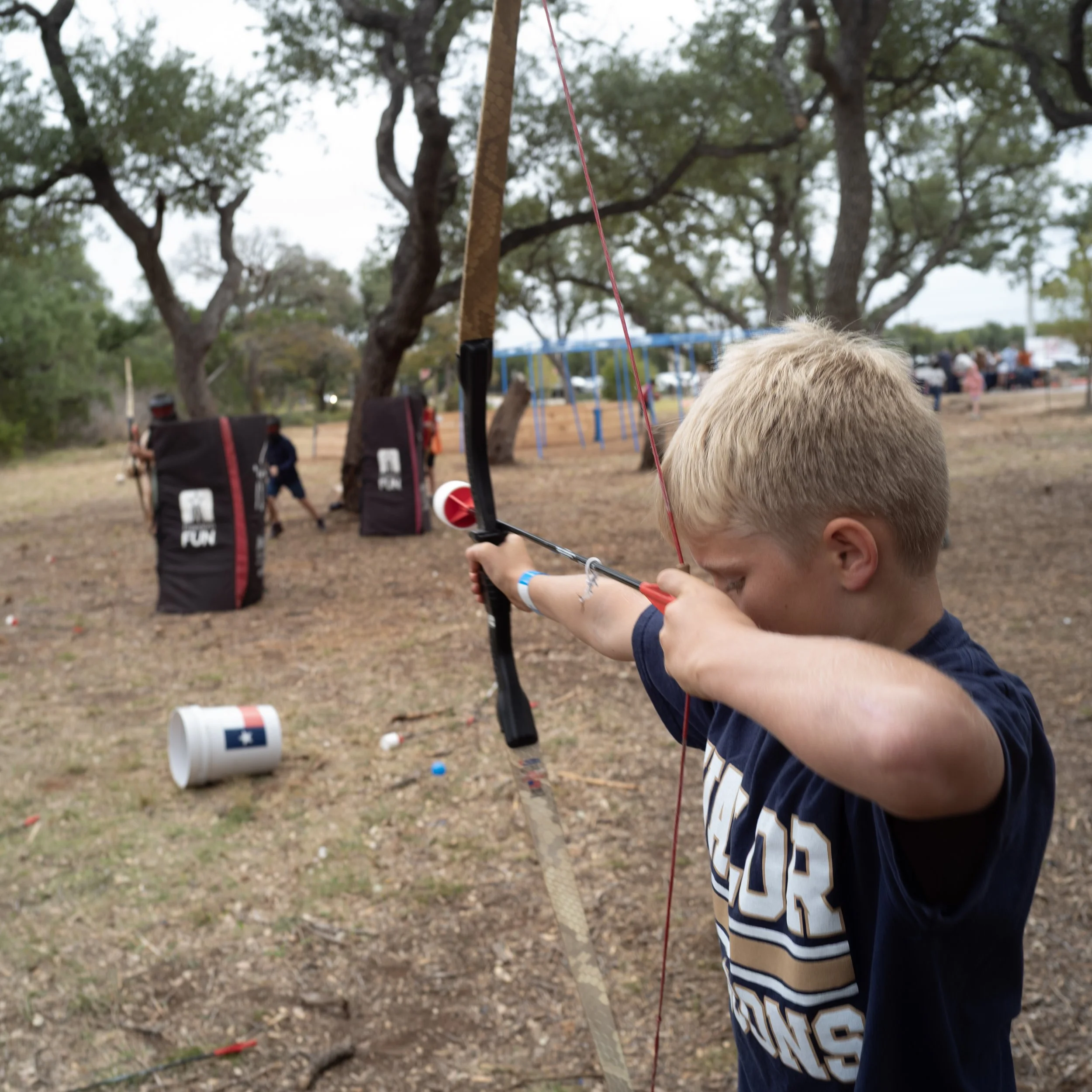 A young boy aiming a bow and arrow outdoors in a park or playground area, with trees and people in the background, and a child’s face partially visible in the foreground.
