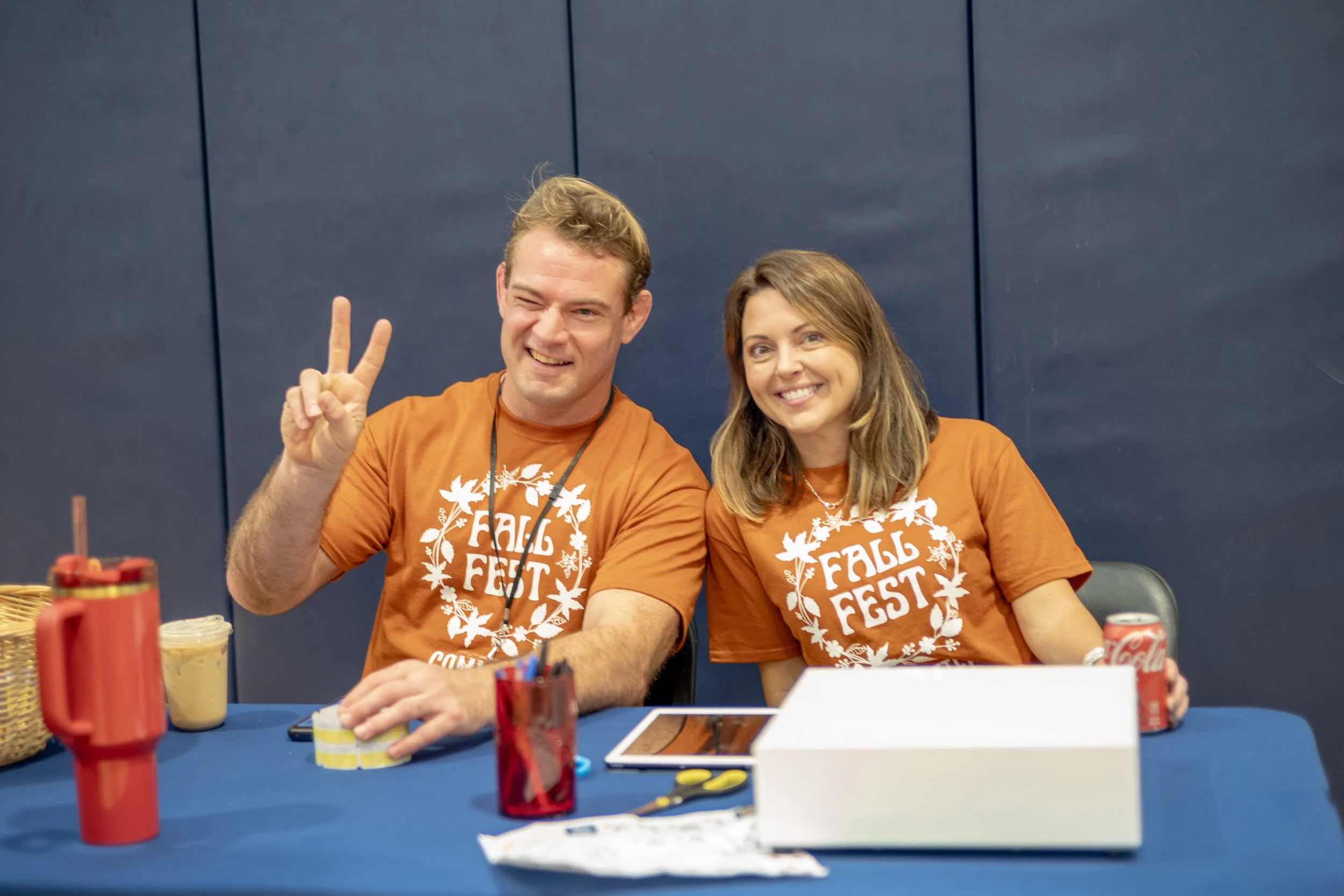Two smiling people at a table wearing matching orange t-shirts with 'Fall Fest' written on them, seated against a blue wall, with various objects like a red thermos, snack containers, and a tablet on the table.