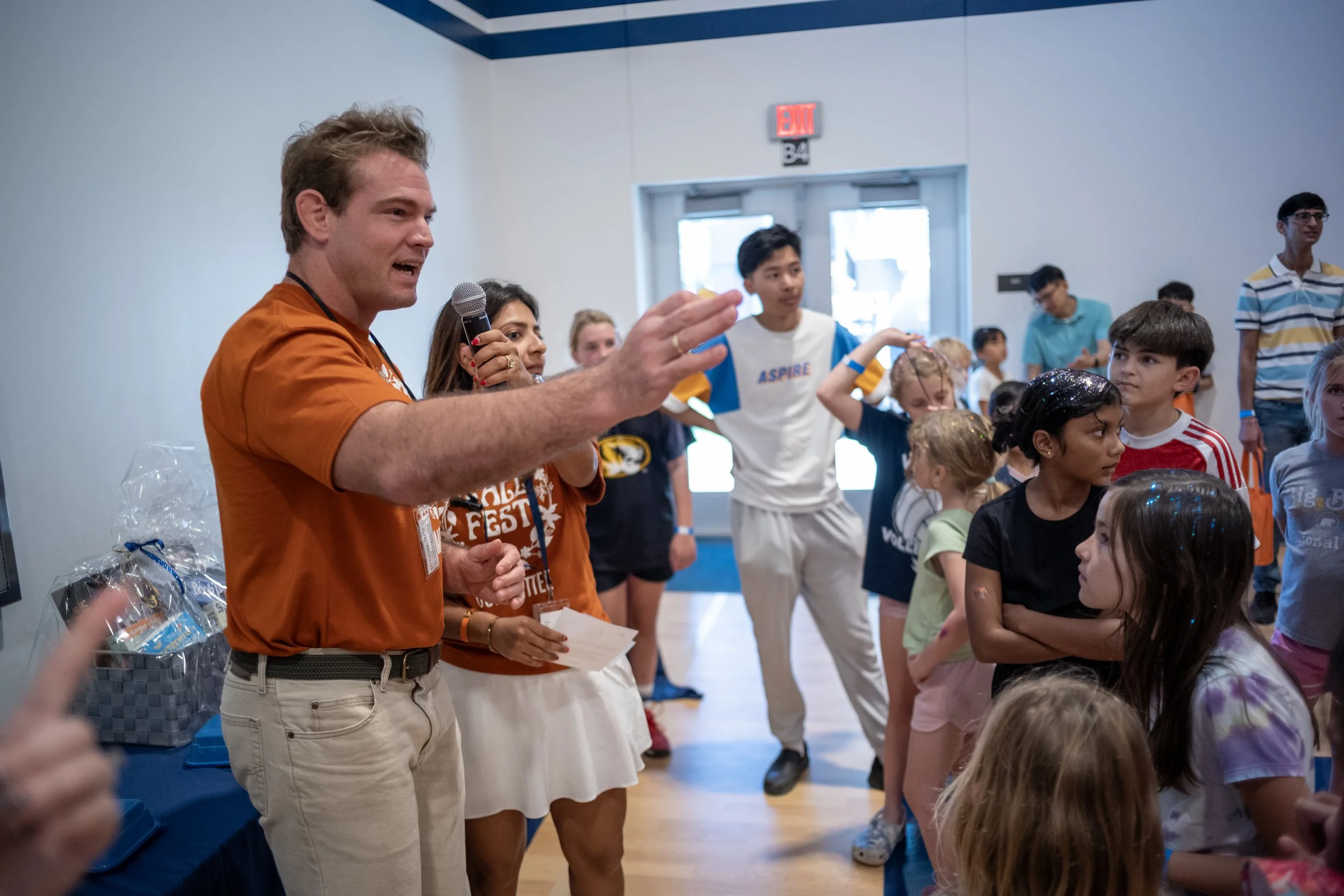 A man with blonde hair and a woman with dark hair, both wearing orange shirts, are speaking to a group of children and teenagers in a gym or cafeteria setting. The man is holding a microphone and gesturing with his hand. The children are listening at