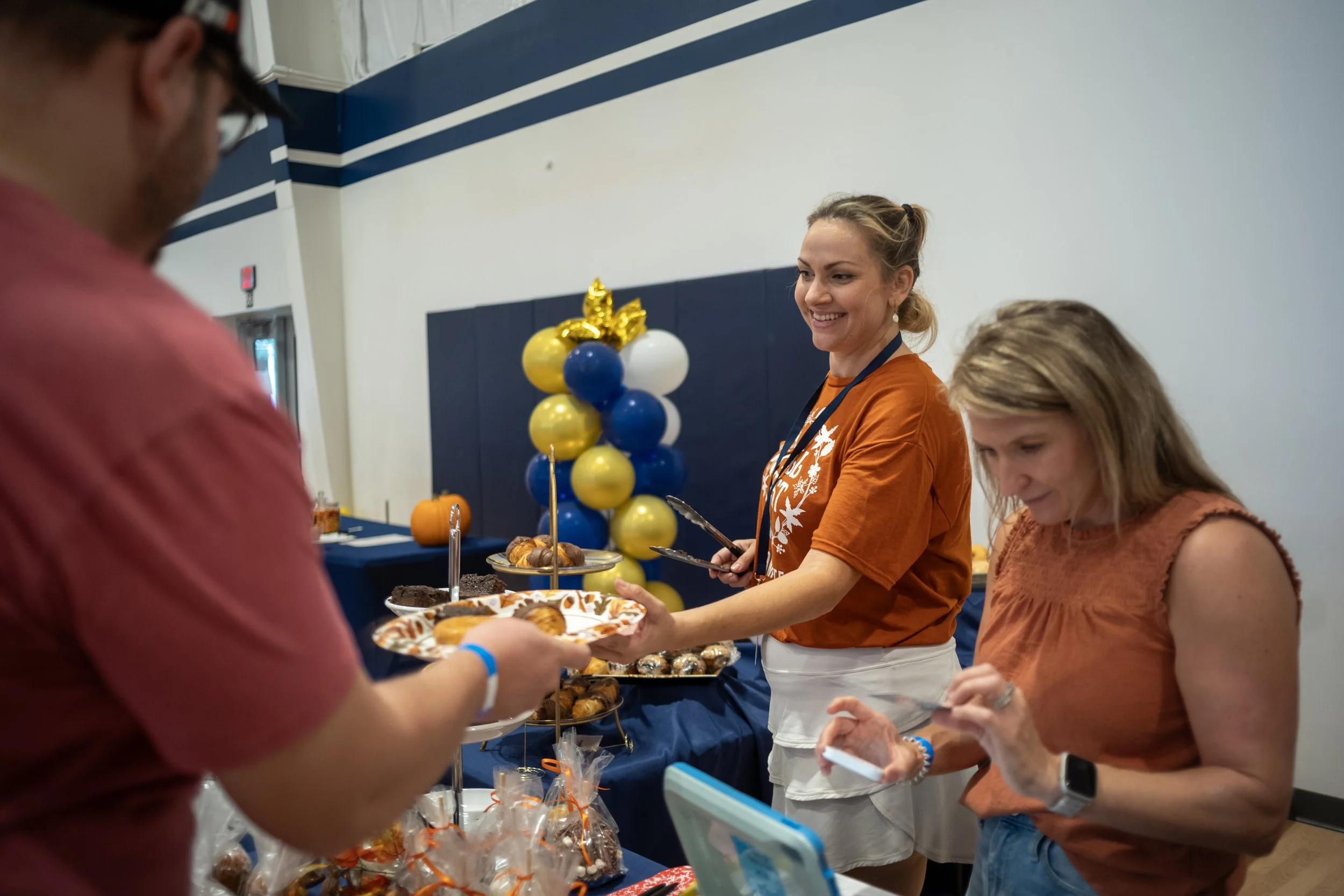 People standing in line at a buffet table with desserts, decorated with balloons, in a large indoor space.