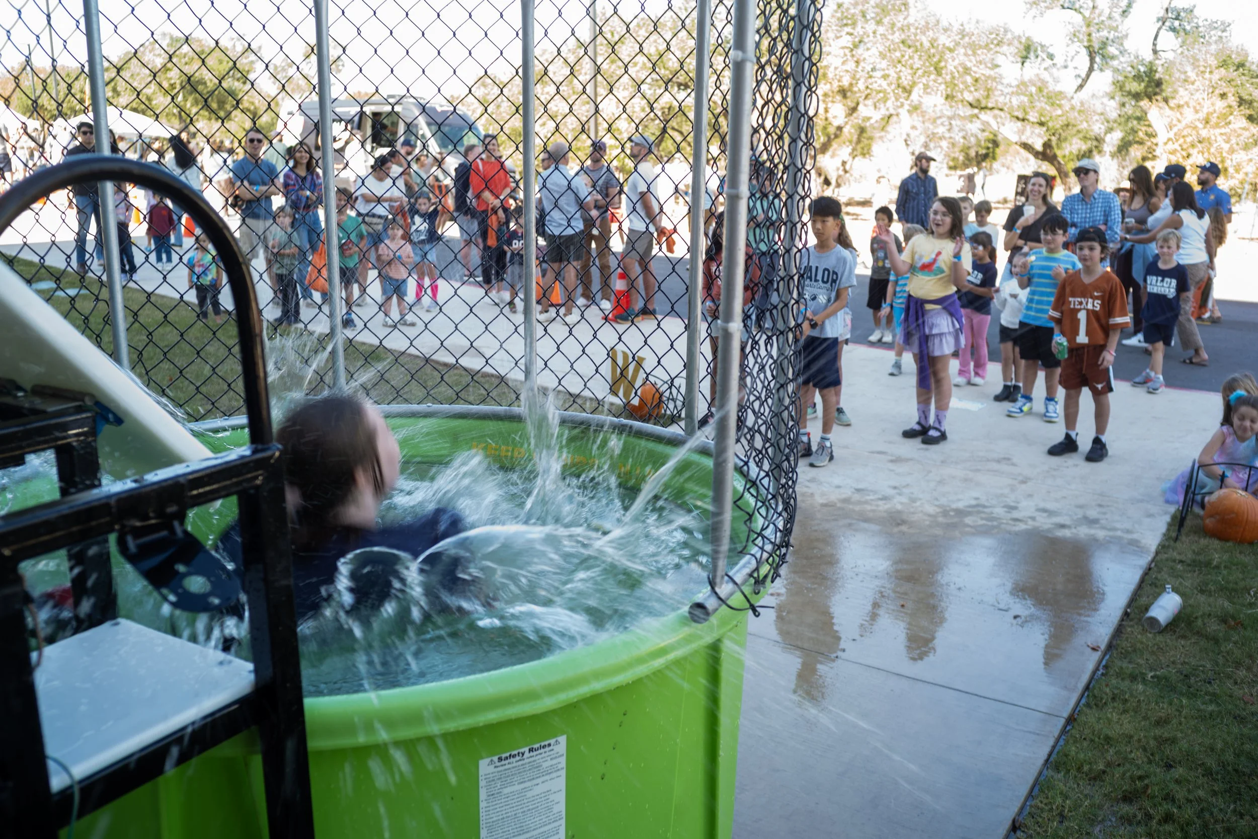 Children and adults waiting in line at a water-themed carnival game on a sunny day, seen through a chain-link fence, with trees and clear sky in the background.