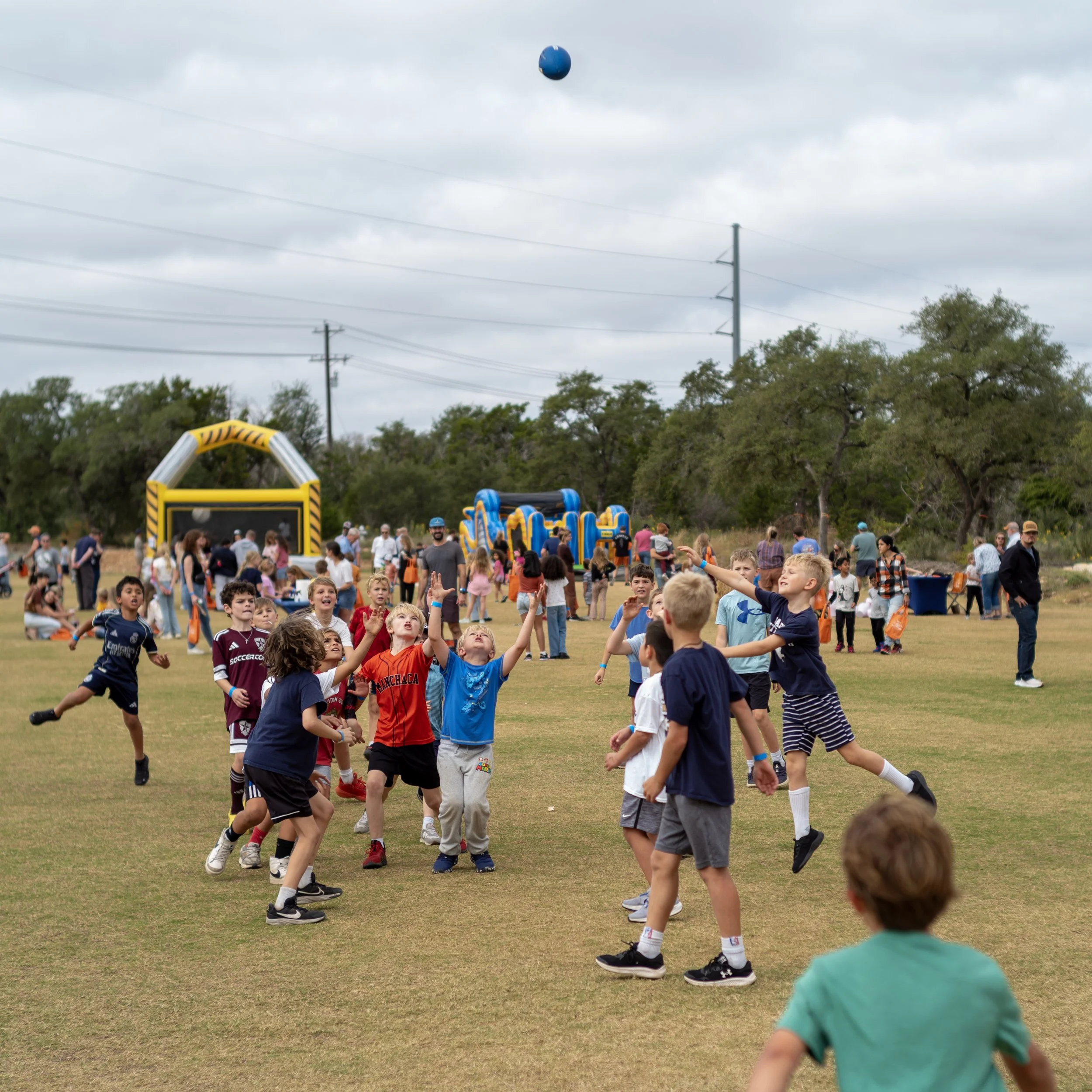 Many children running and playing outdoors on a field during a cloudy day, with amusement rides and trees in the background.