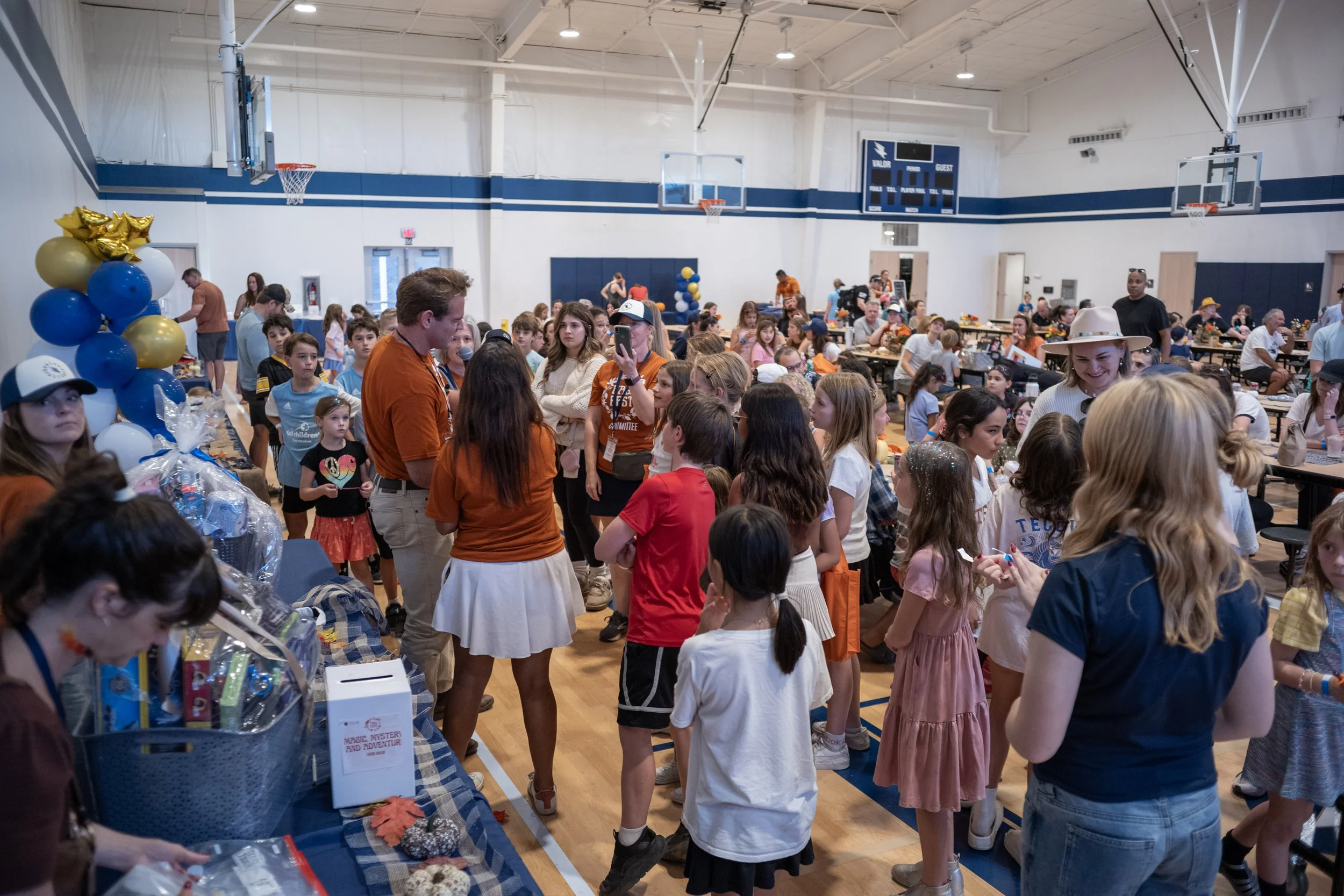 A crowded indoor event in a gymnasium filled with children and adults, with balloons and tables, some people wearing themed clothing, and a man with a microphone speaking to the crowd.