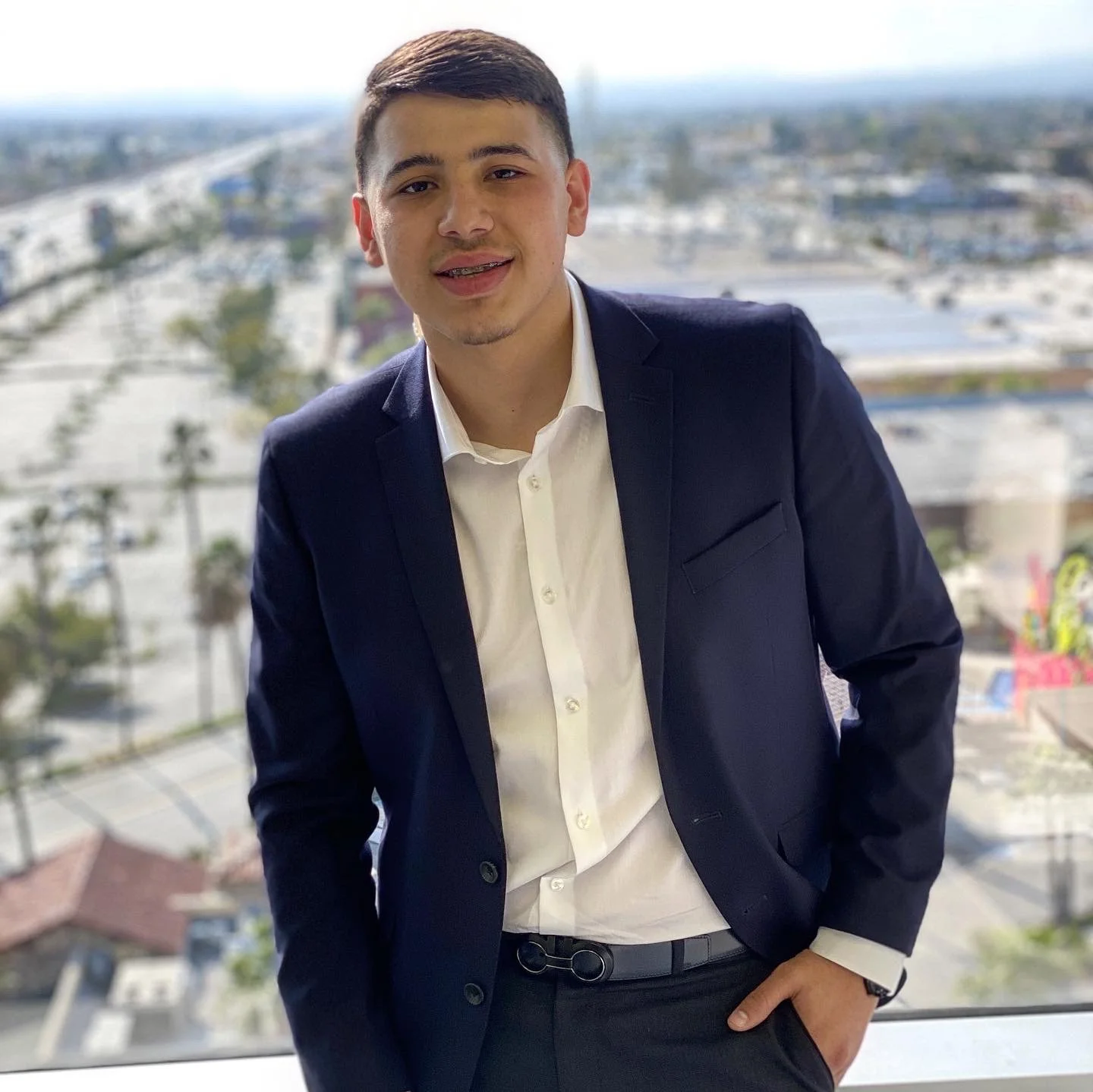 Young man in a suit smiling, standing by a window with a cityscape view of buildings, trees, and a parking lot in the background.