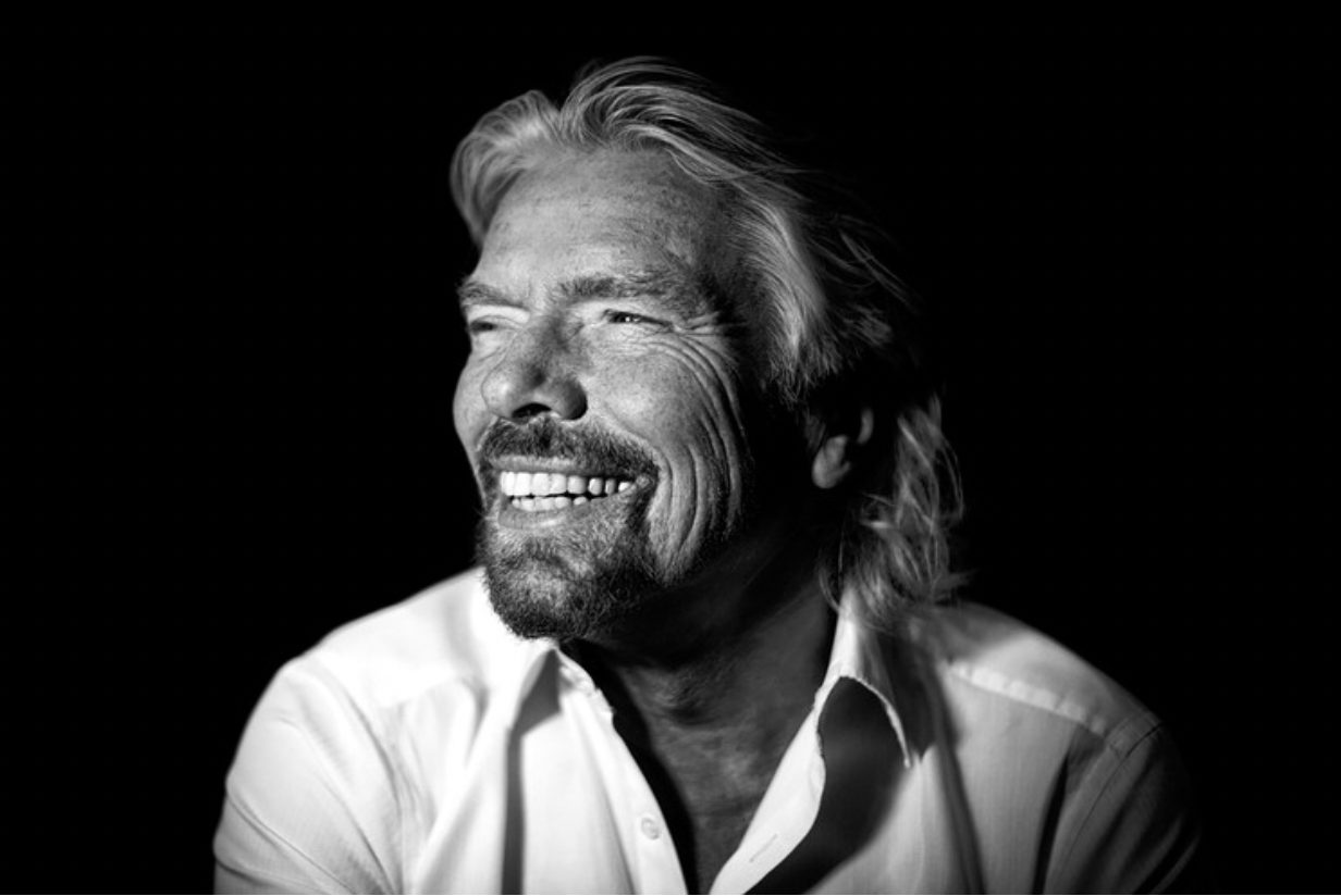 Black and white portrait of a smiling middle-aged man with long hair and a beard, wearing a white shirt, against a dark background.