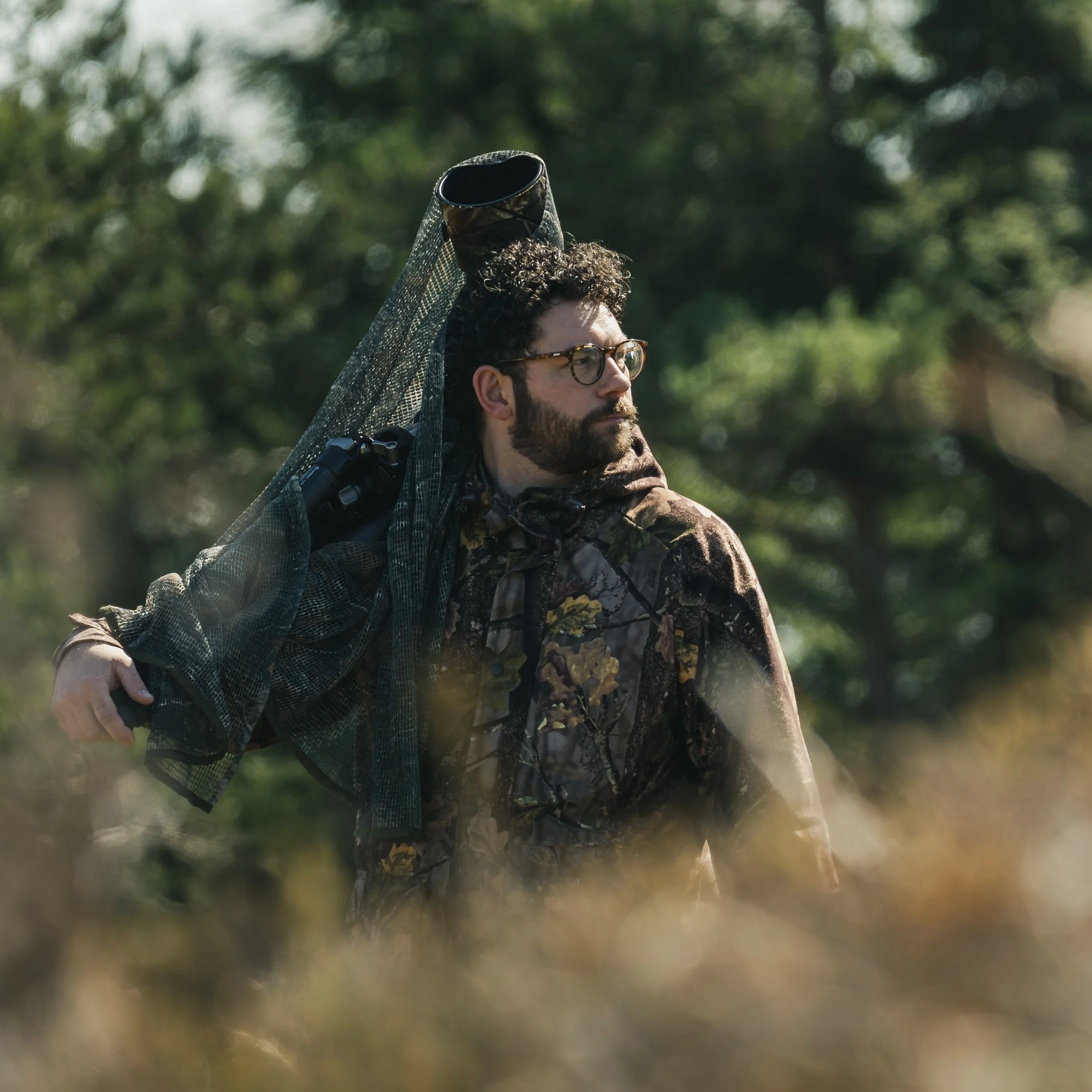 A man with glasses and a beard wearing camouflage clothing, carrying a Sony Wildlife Camera over his shoulder, stands outdoors in a green, wooded area.