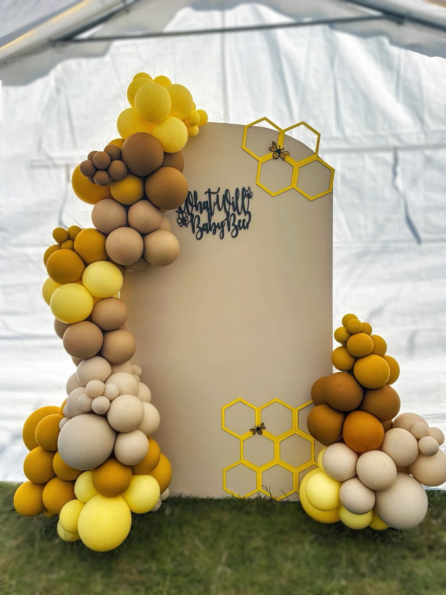 Decorative backdrop with a cluster of balloons in shades of yellow, brown, and white arranged around a white board, featuring a sign that reads 'What Will Baby Bee' and yellow honeycomb-shaped frames with bee decorations.