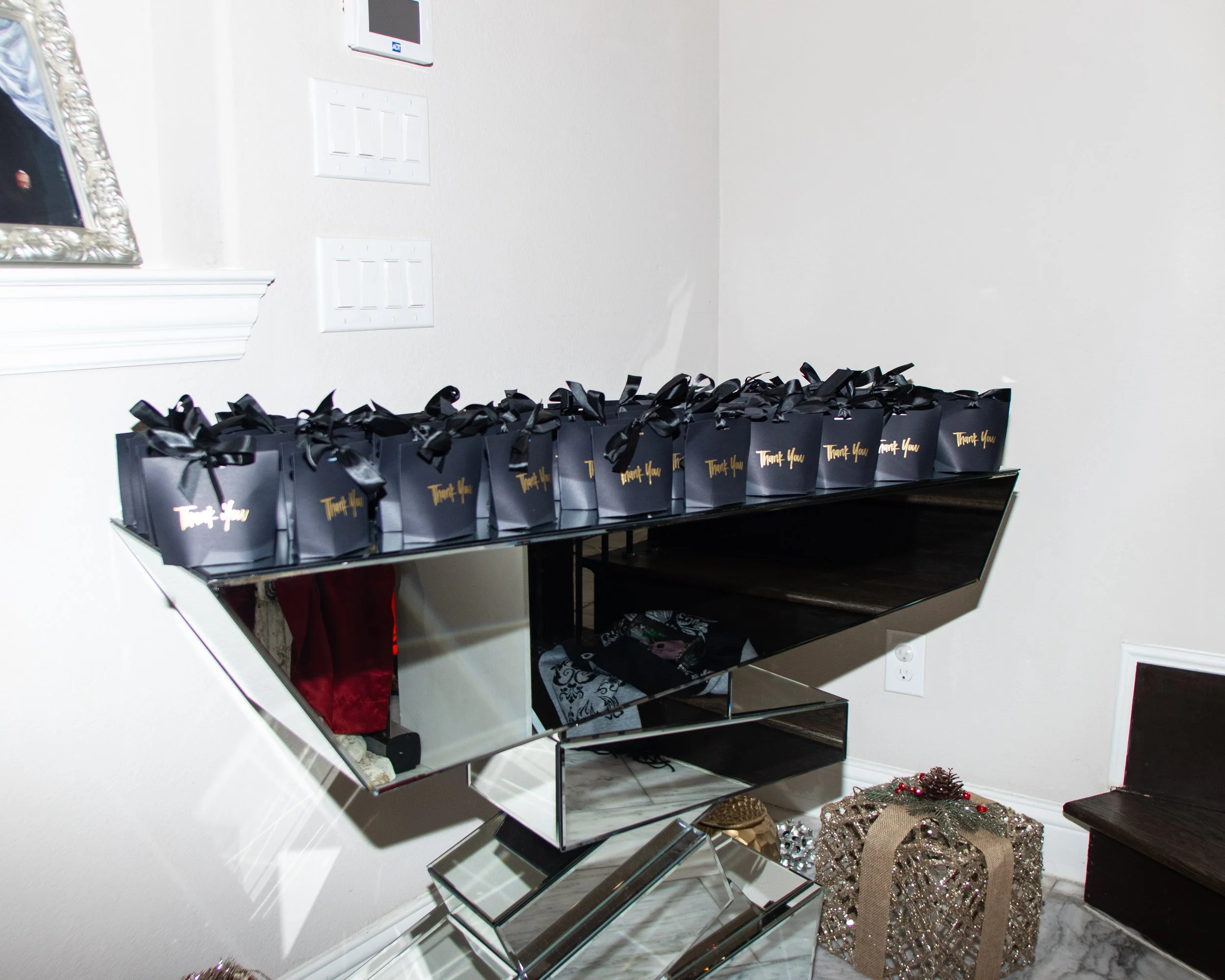 A row of black gift bags with gold 'Thank You' written on them, lined up on a black glass table, and a decorative gift box on the floor nearby.