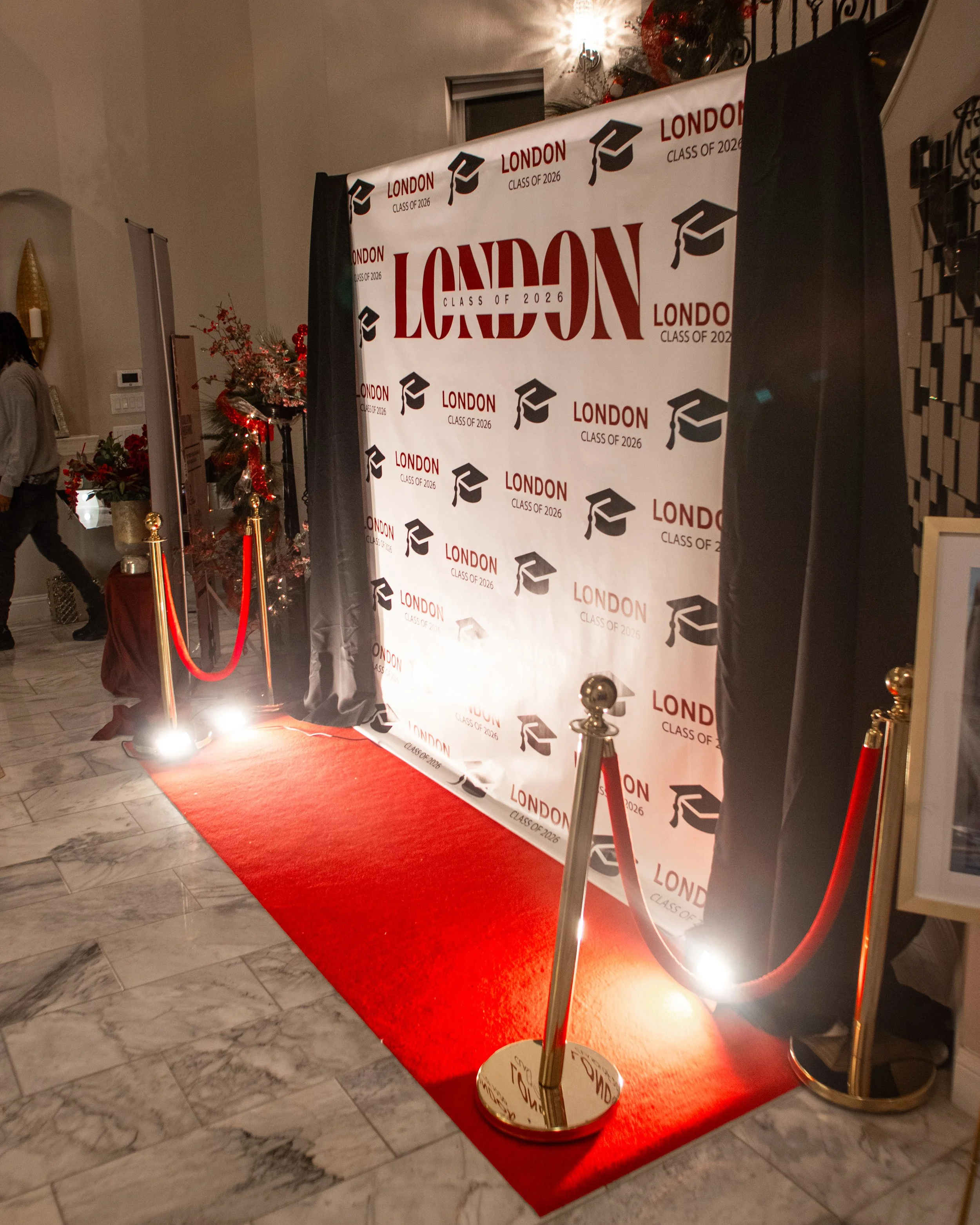 Red carpet event backdrop with black curtains, decorated with Christmas ornaments, next to a Christmas tree. The backdrop has the text 'LONDON CLASS OF 2026' with graduation cap icons.