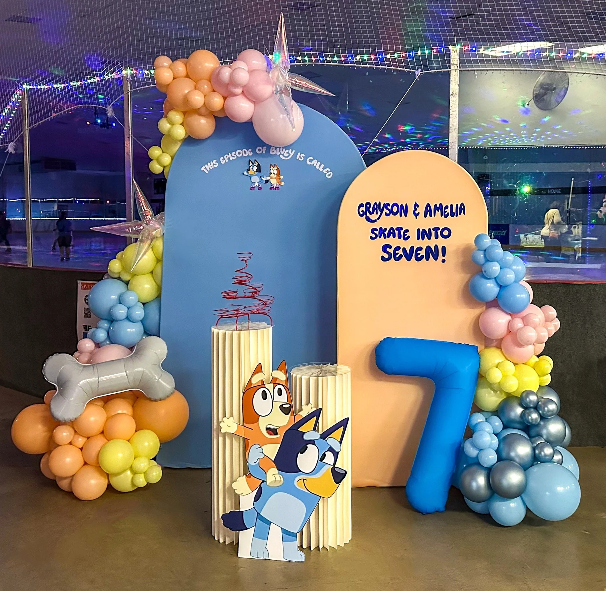 Decorative birthday party display for a 7th birthday, featuring balloons, cartoon character cutouts of Bluey and Bingo, a large blue number 7, and birthday messages, with a skating rink in the background.