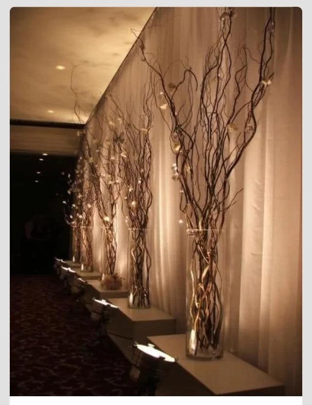 Tall vases filled with twisted decorative branches and string lights, displayed along a wall with curtains in a dimly lit corridor.