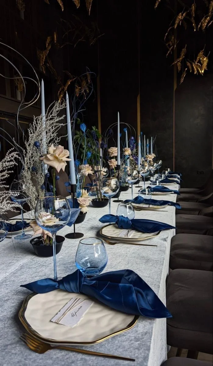 Elegant dining table set for a special event, with cream-colored plates, blue cloth napkins tied in bows, clear wine glasses, and tall candles. The table has a floral centerpiece with white and blue flowers and decorative elements, and the setting is in a dimly lit room with dark walls.