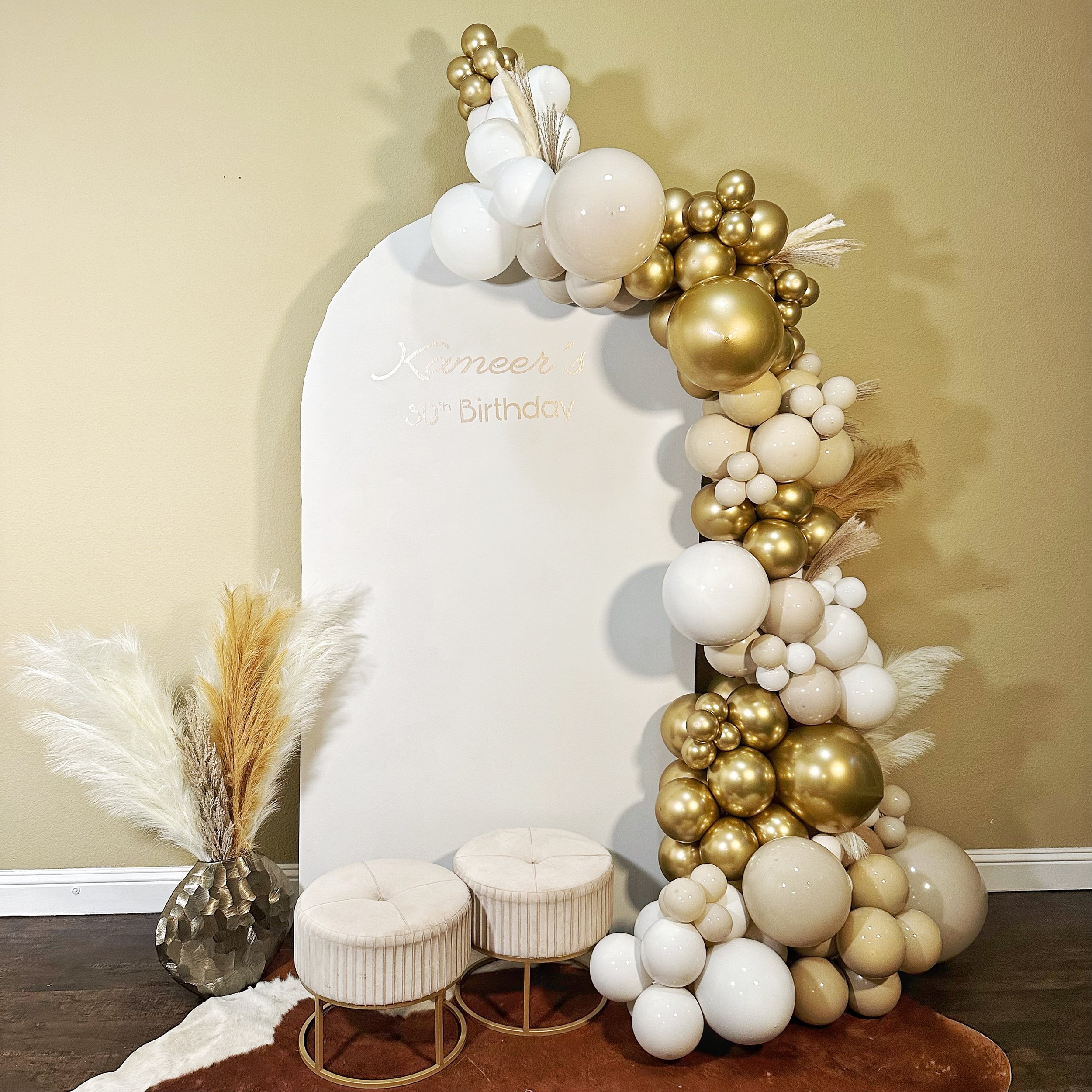 Decorative birthday party backdrop with white and gold balloons arranged in an arch, a white oval board with gold lettering reading "Kameer's 3rd Birthday", a brown vase with pampas grass, and two round upholstered stools in front, set on a white and