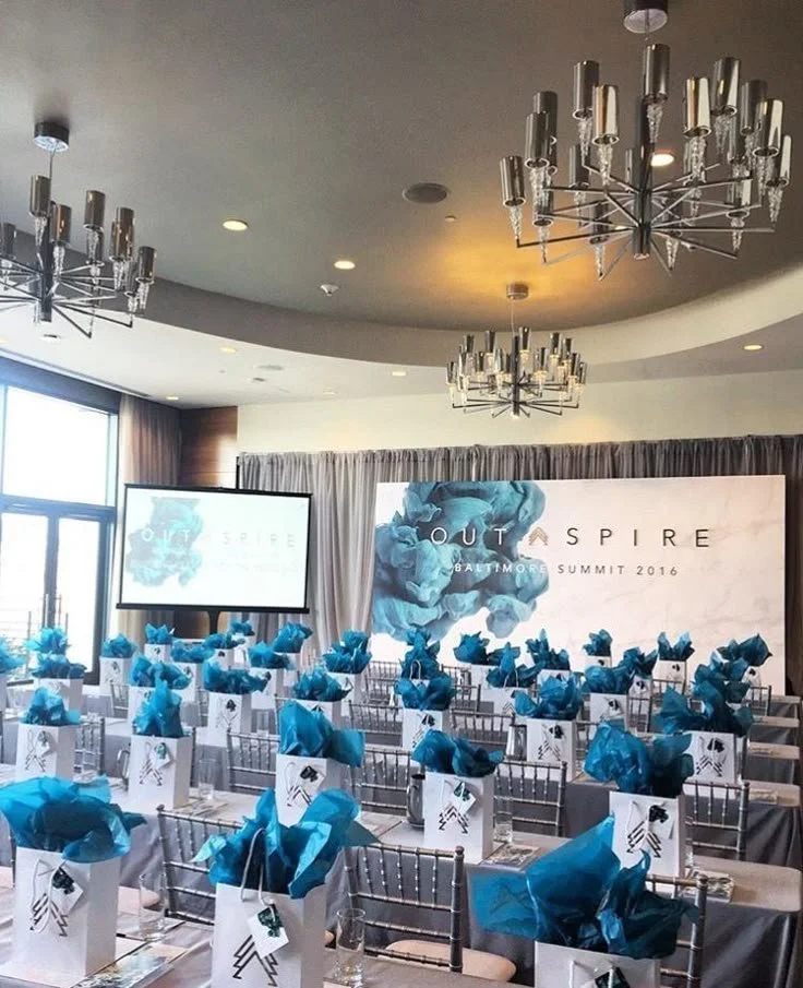 Conference room set up for an event, with chairs, gift bags with blue tissue paper, and a large screen displaying a logo and the words 'OUT + SPIRE Baltimore Summit 2016.'