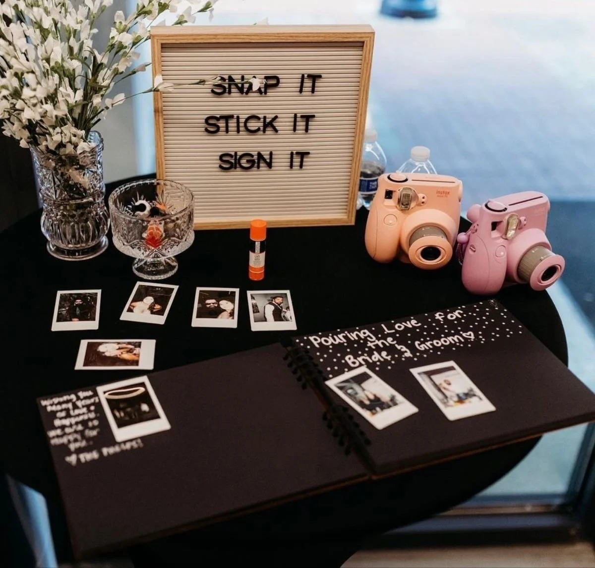 Table decorated with a sign that reads "SNAP IT, STICK IT, SIGN IT," two pink instant cameras, a clipboard recording wedding moments, several Polaroid photographs, a glass vase with white flowers, a small glass bowl, two bottles of water, a glue stic