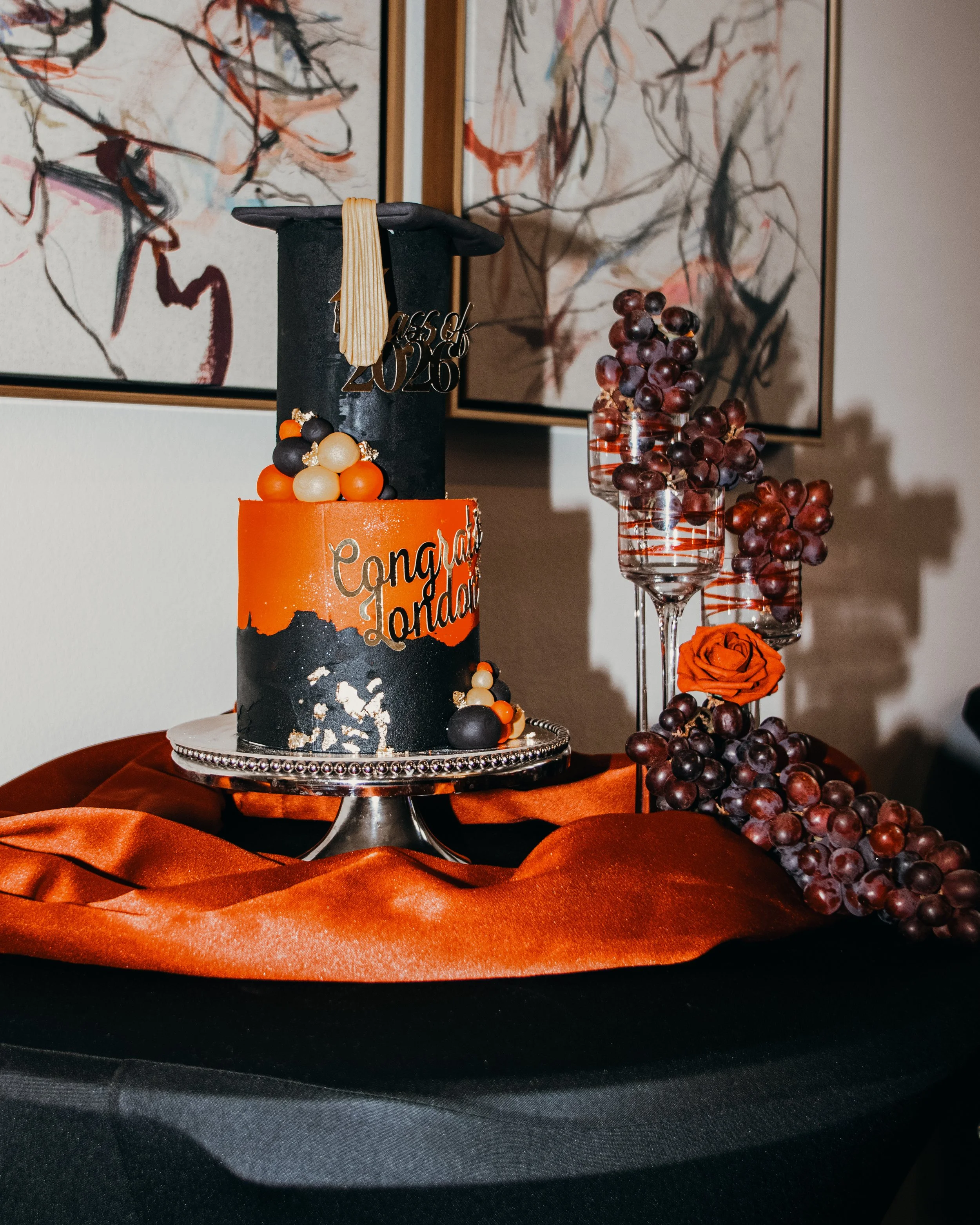 A graduation cap and cake with decorations, and two glasses filled with grapes, on a table covered with an orange cloth.