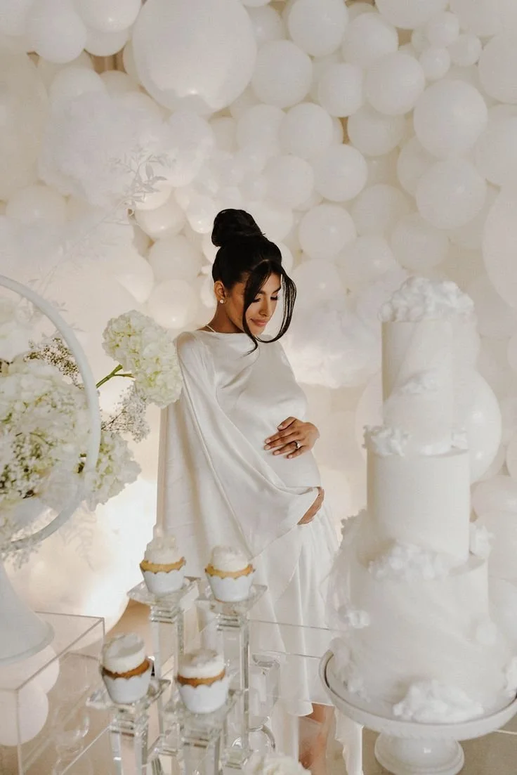 A pregnant woman in white dress at a white-themed celebration with cake, cupcakes, and white balloons.