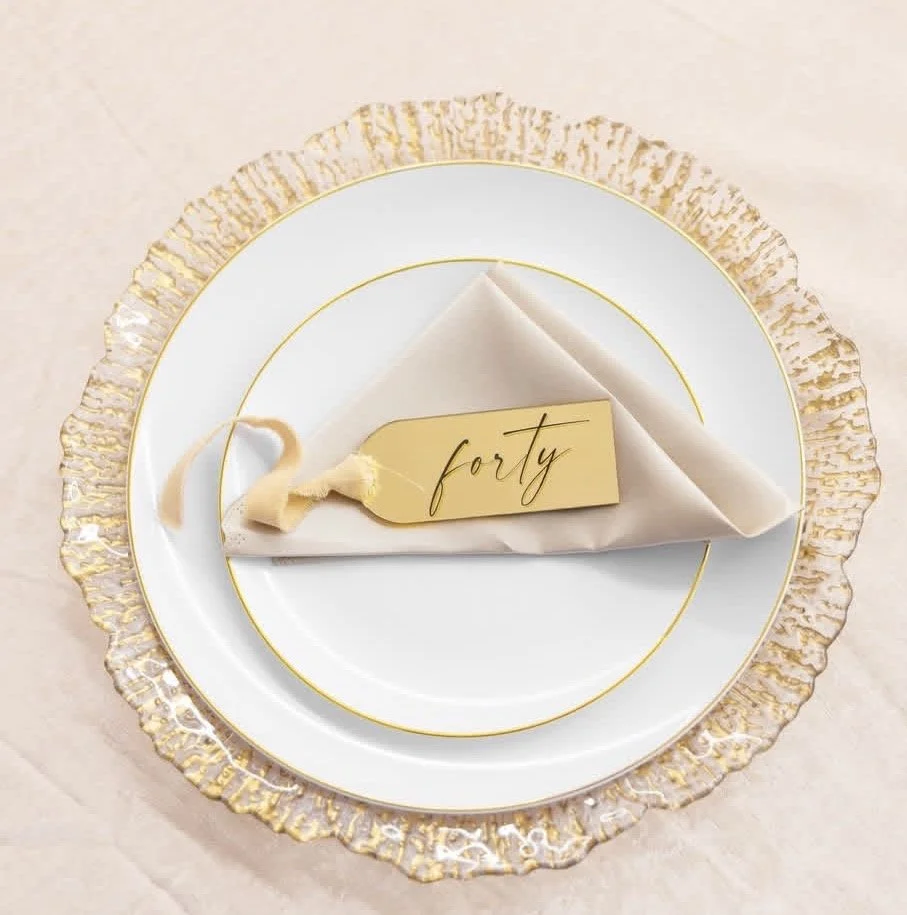 Elegant table setting with a gold-rimmed plate and a folded white napkin, decorated with a small yellow tag that reads 'forty'.