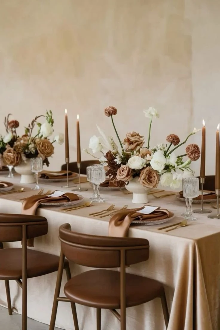 Elegant dining table with beige tablecloth, floral centerpieces, brown napkins, gold utensils, and candle holders with candles.