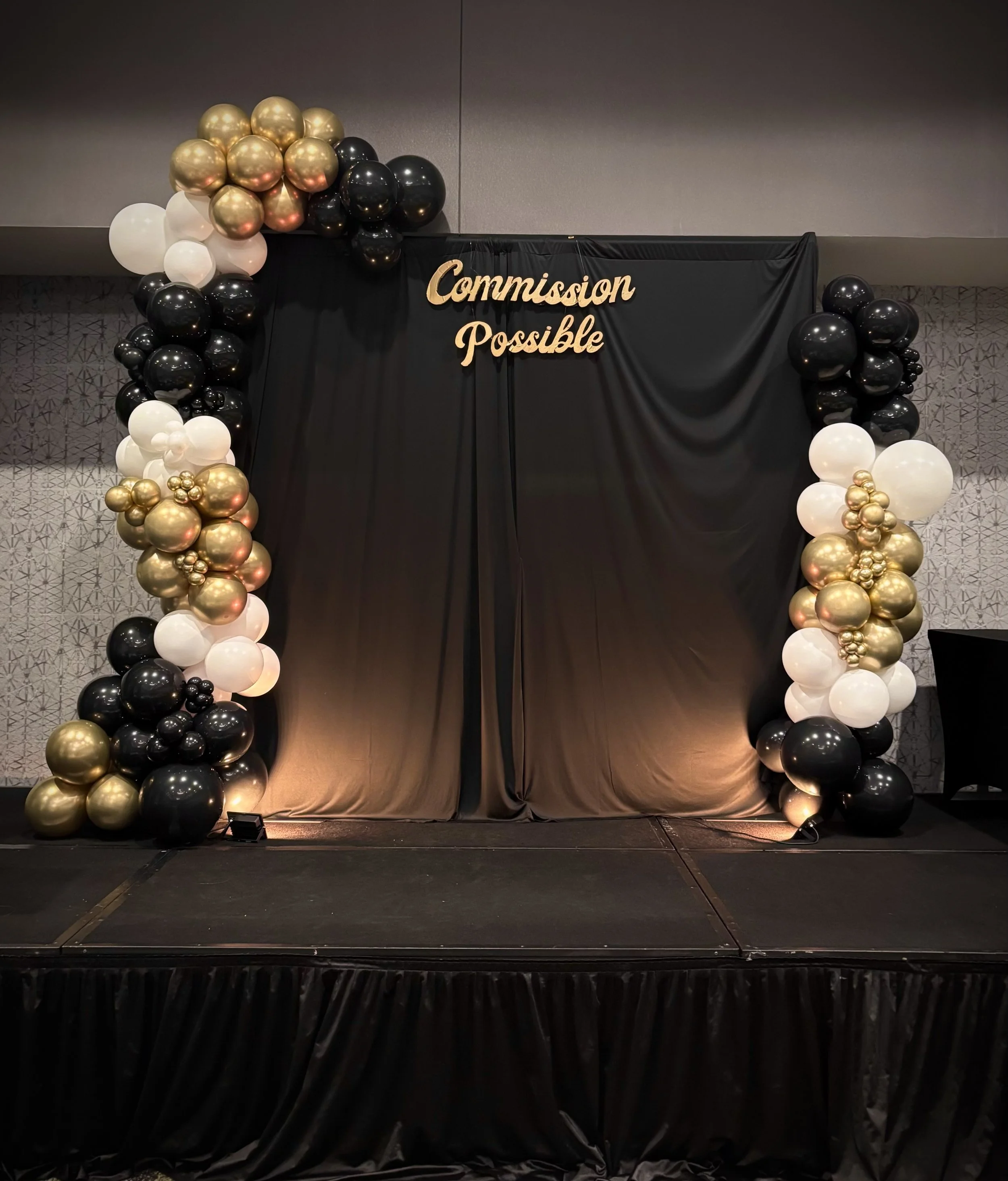 Stage decorated with black curtains, gold, black, and white balloons, and a sign that says "Commission Possible."
