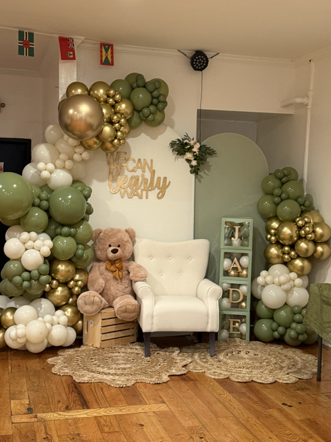 Decorative backdrop with balloons, a teddy bear, a white chair, and decorated blocks spelling 'JADEN', likely for a celebration or photo backdrop.