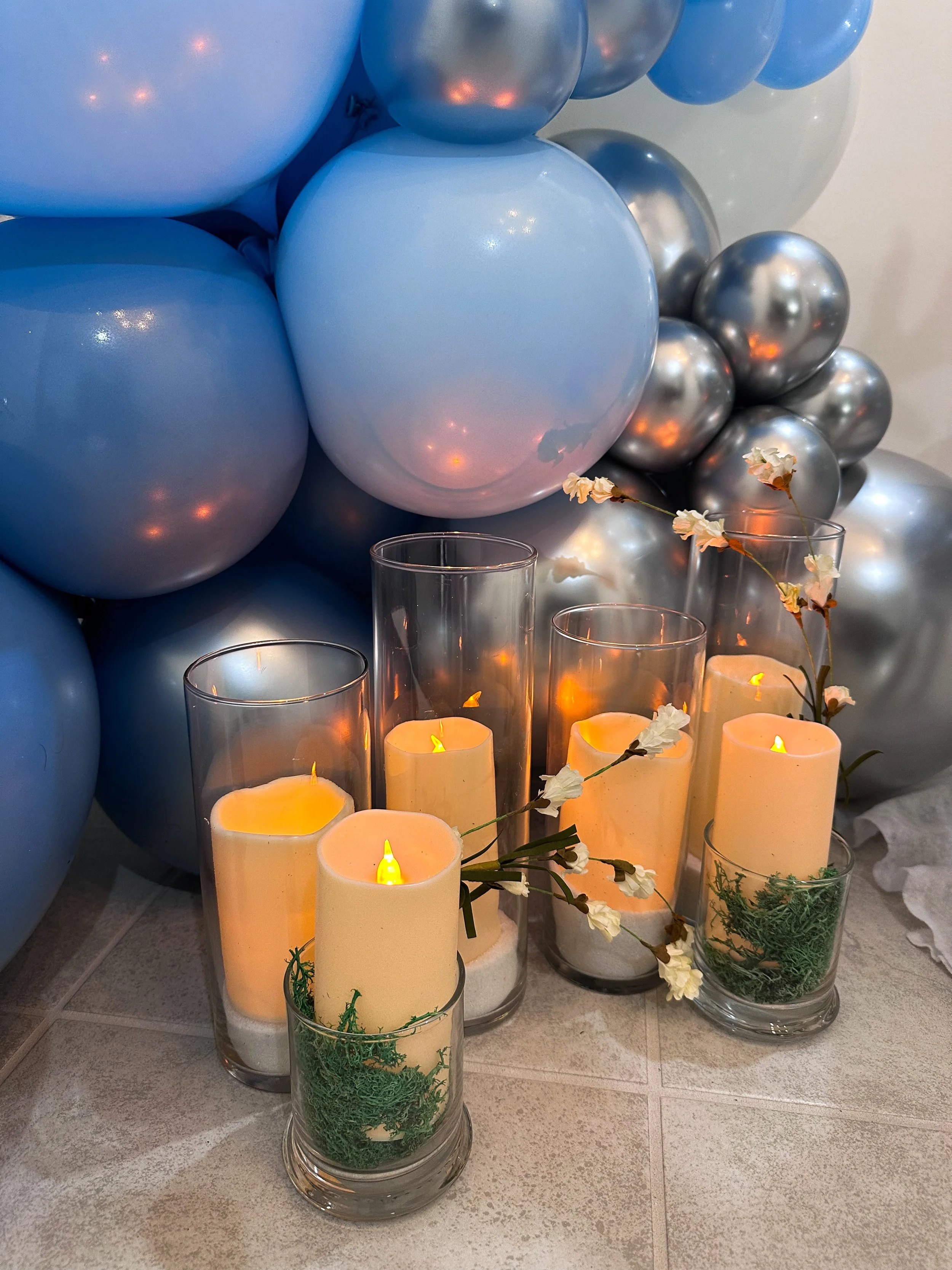 A decorative arrangement of blue and silver balloons with lit candles in glass holders, accented with white flowers and greenery.