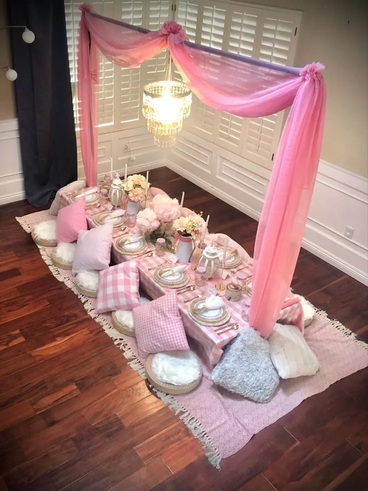 A low, pink and white picnic-style table set up for a gathering, decorated with pink flowers, candles, and tableware. Cushions surround the floor for seating. A pink canopy draped over the table and a chandelier hang above.