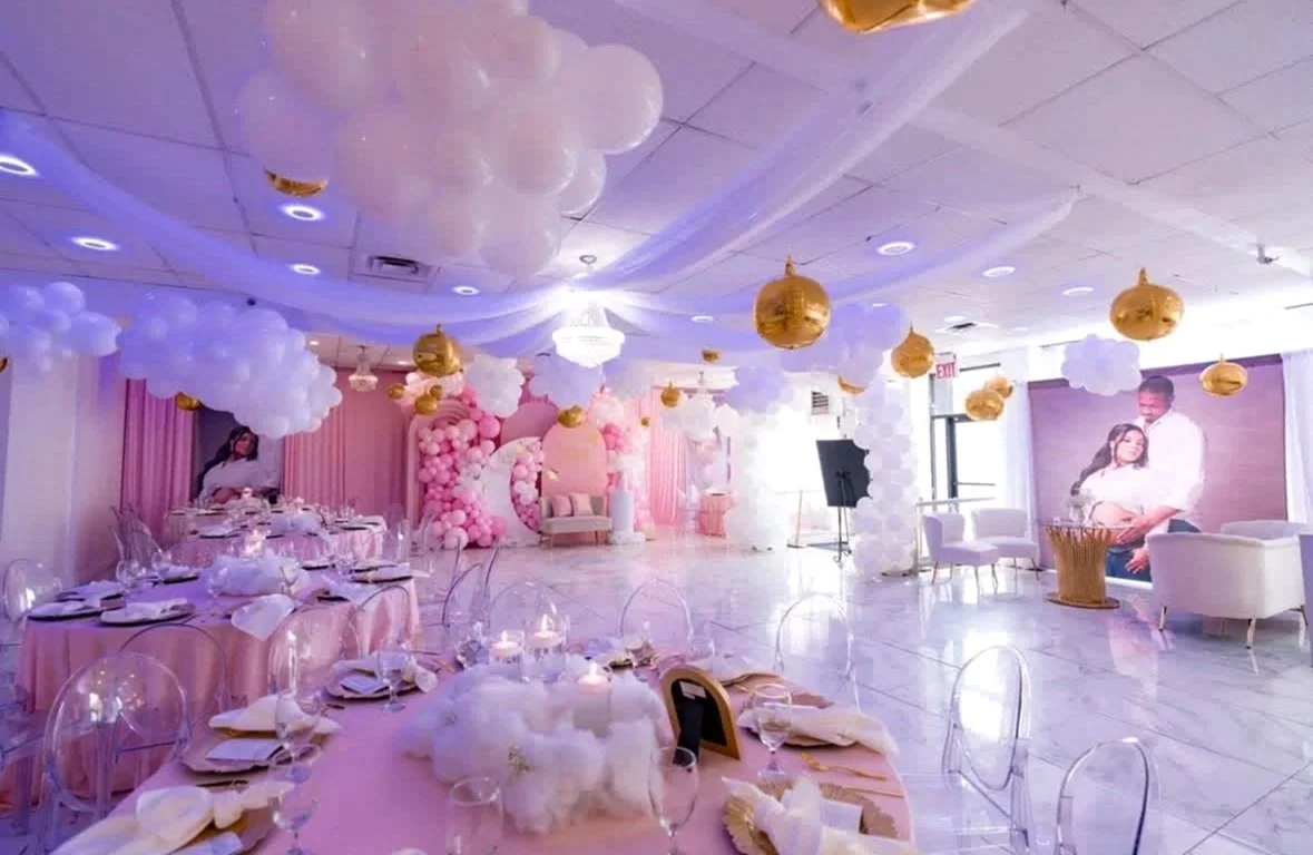 Decorated event space with pink and white balloons, clouds, and gold accents, hosting a celebration with round tables, chairs, and a large screen displaying a couple.
