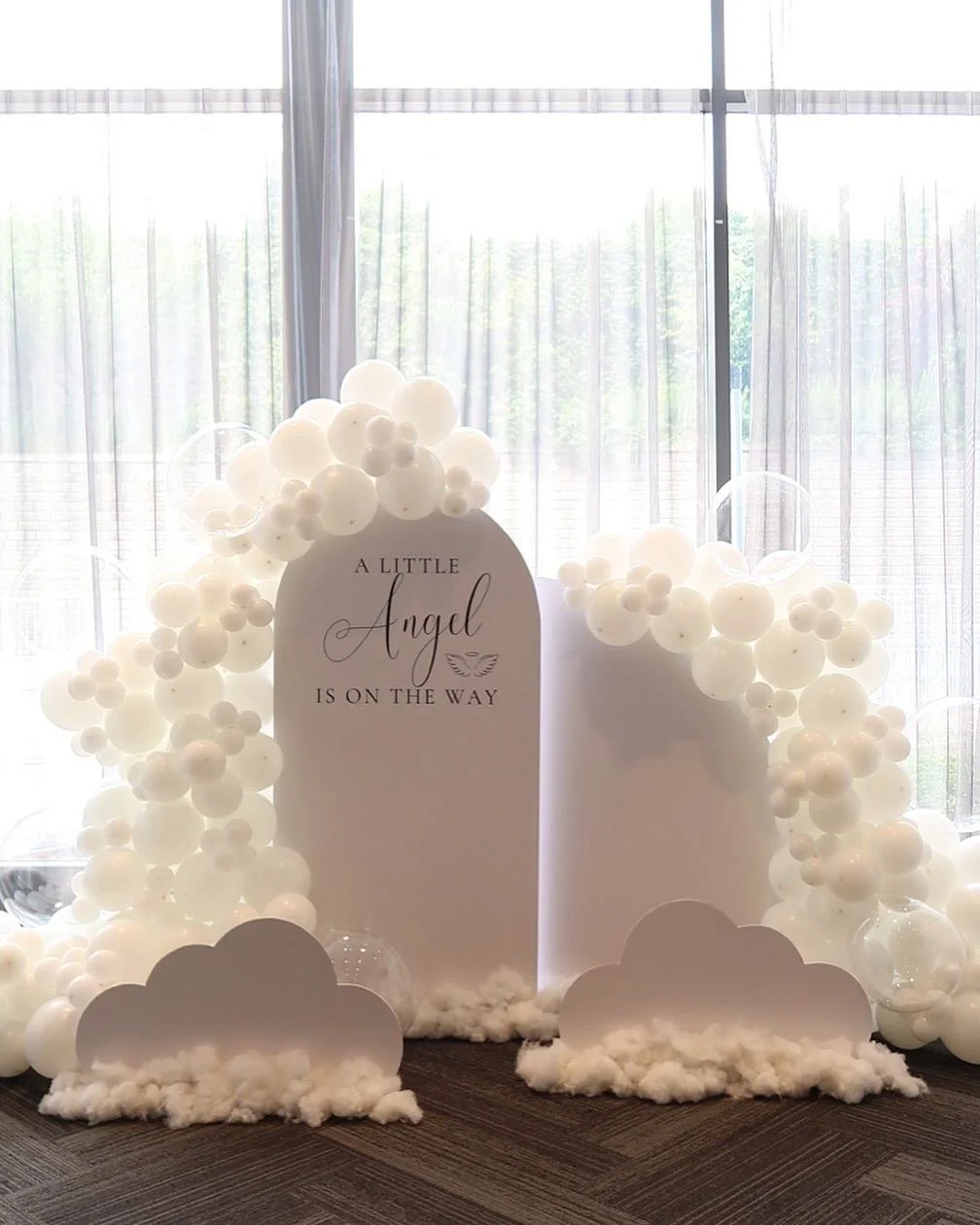 A decorative baby shower display with white balloons, cloud cutouts, and a sign that says "A little Angel is on the way" with a small angel wing illustration.