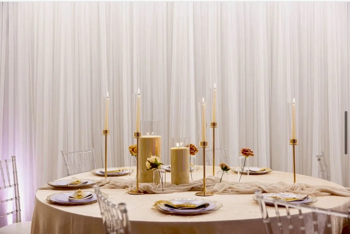 Elegant dining table setup with tall candles, candles of varying heights, white floral accents, and transparent chairs, set against a cream-colored curtain backdrop.