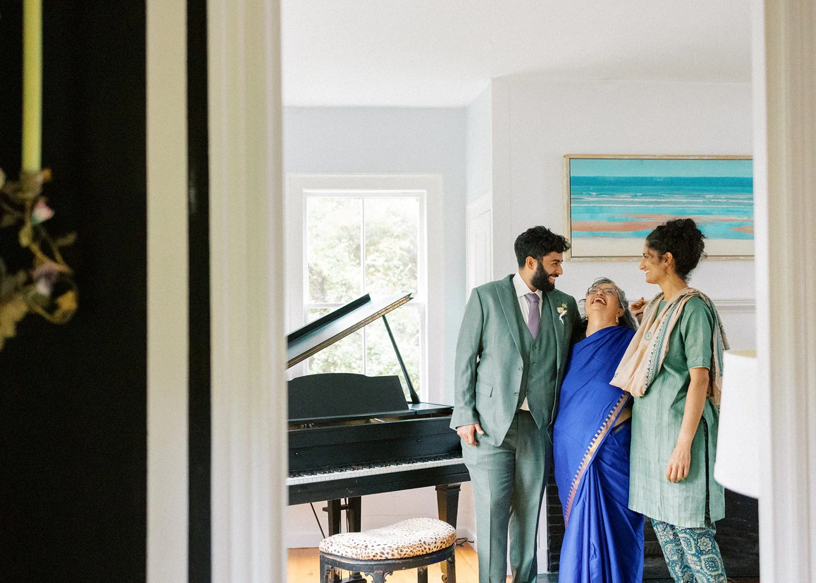 A joyful moment with three people smiling at each other in a living room, with a black grand piano in the background and an abstract seascape painting on the wall.