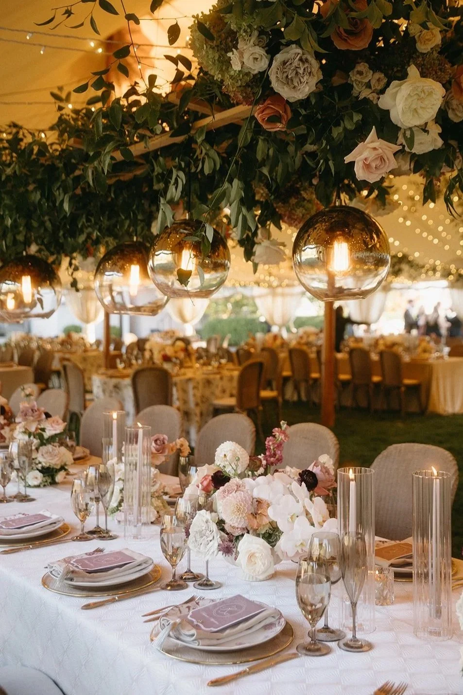 Elegant event setup with decorated long banquet tables, floral centerpieces, candles, and hanging glass globe lights under a canopy of greenery and flowers.