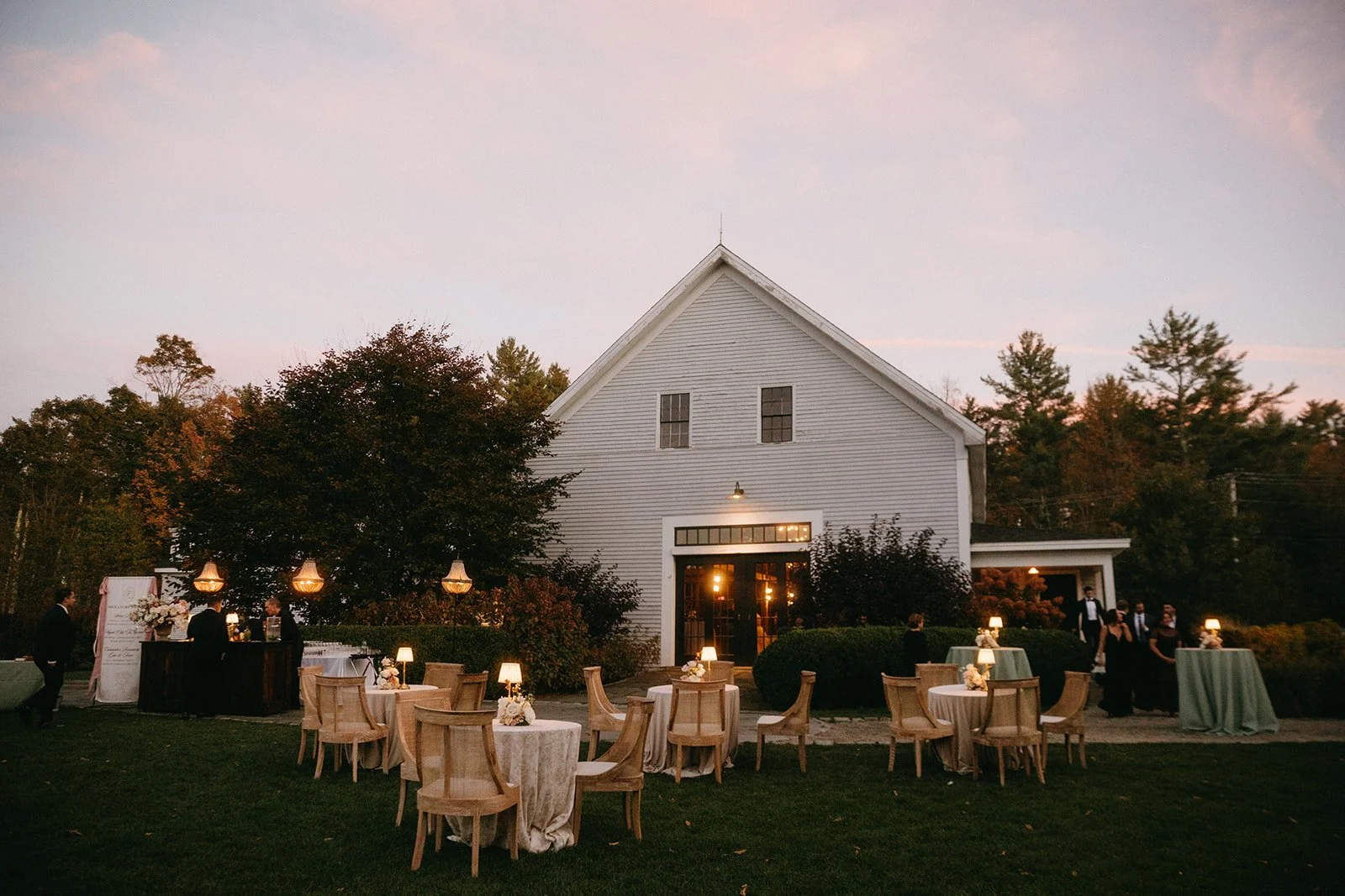 Outdoor evening wedding reception with round tables, lanterns, and a white barn building surrounded by trees.